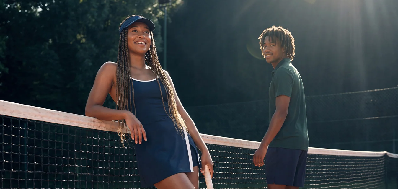 Due giovani stanno su un campo da tennis in terra rossa vicino alla rete, uno appoggiato con una racchetta, luce del sole e alberi sullo sfondo.
