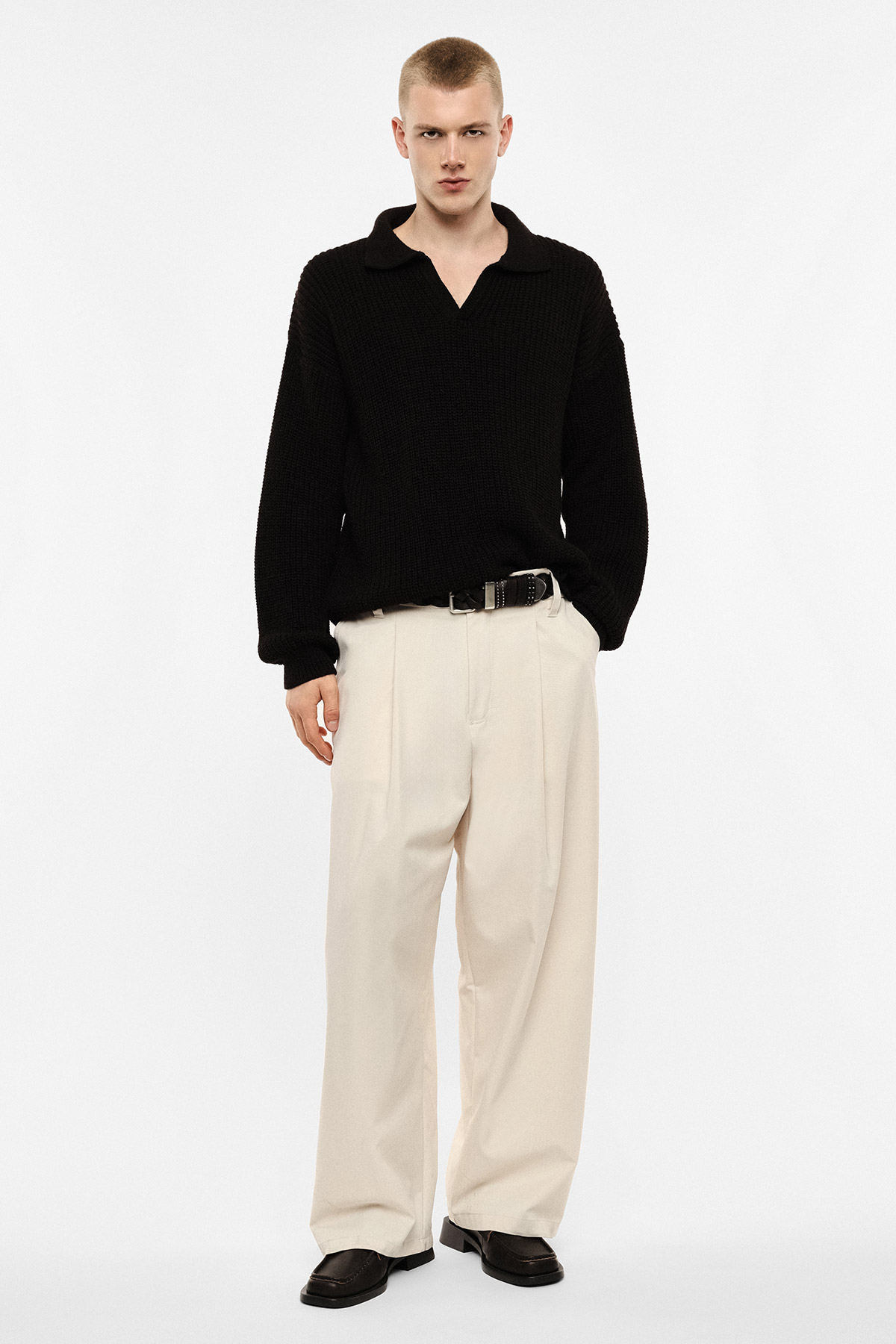 Model wearing black knitted sweater, cream wide-leg trousers and black loafers on white background
