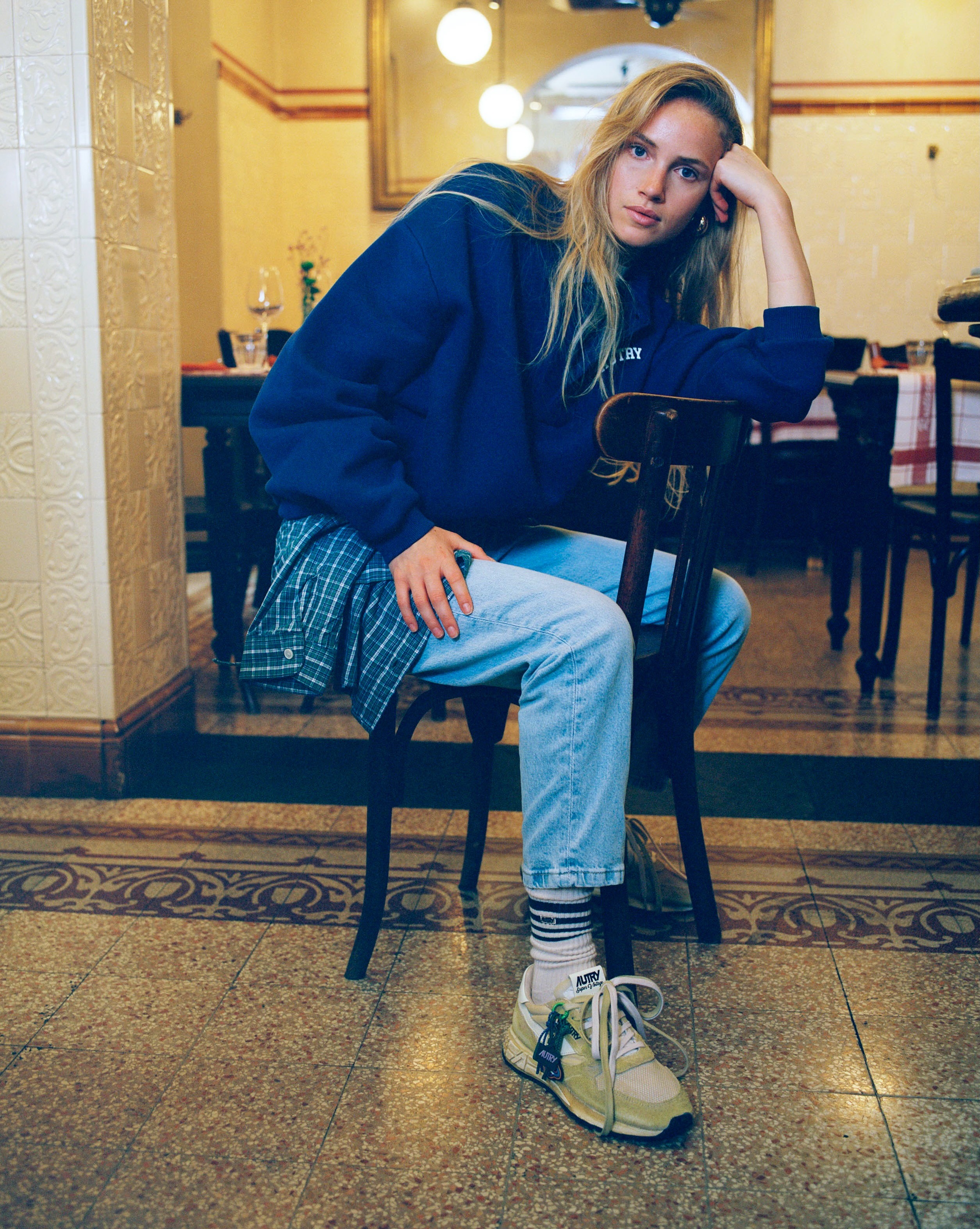 Young woman in blue sweatshirt and jeans sitting on a chair in a cosy café, leaning on hand, wearing trainers