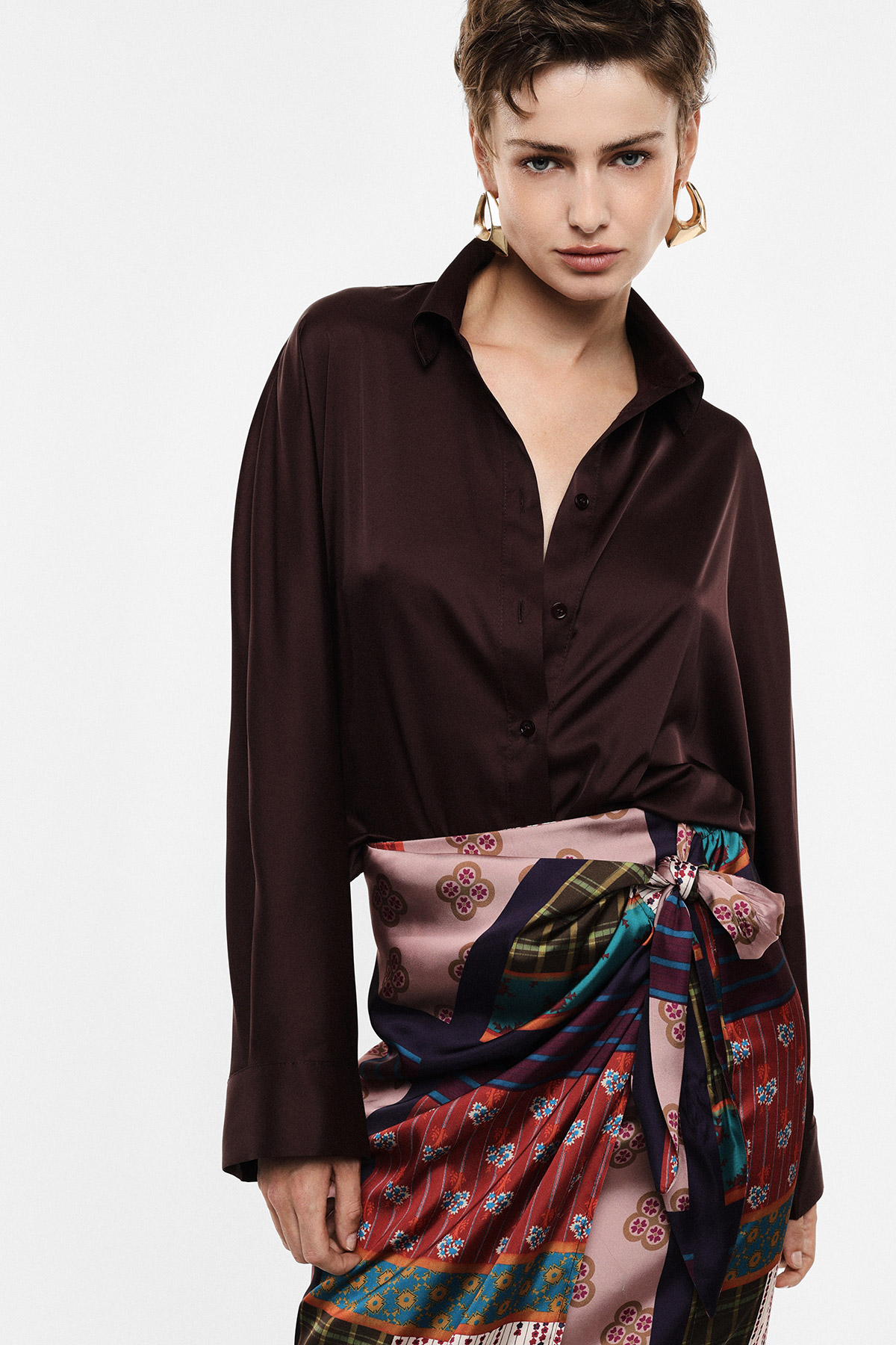 Model wearing dark brown silk blouse and colourful patterned silk wrap skirt tied at the waist, gold hoop earrings