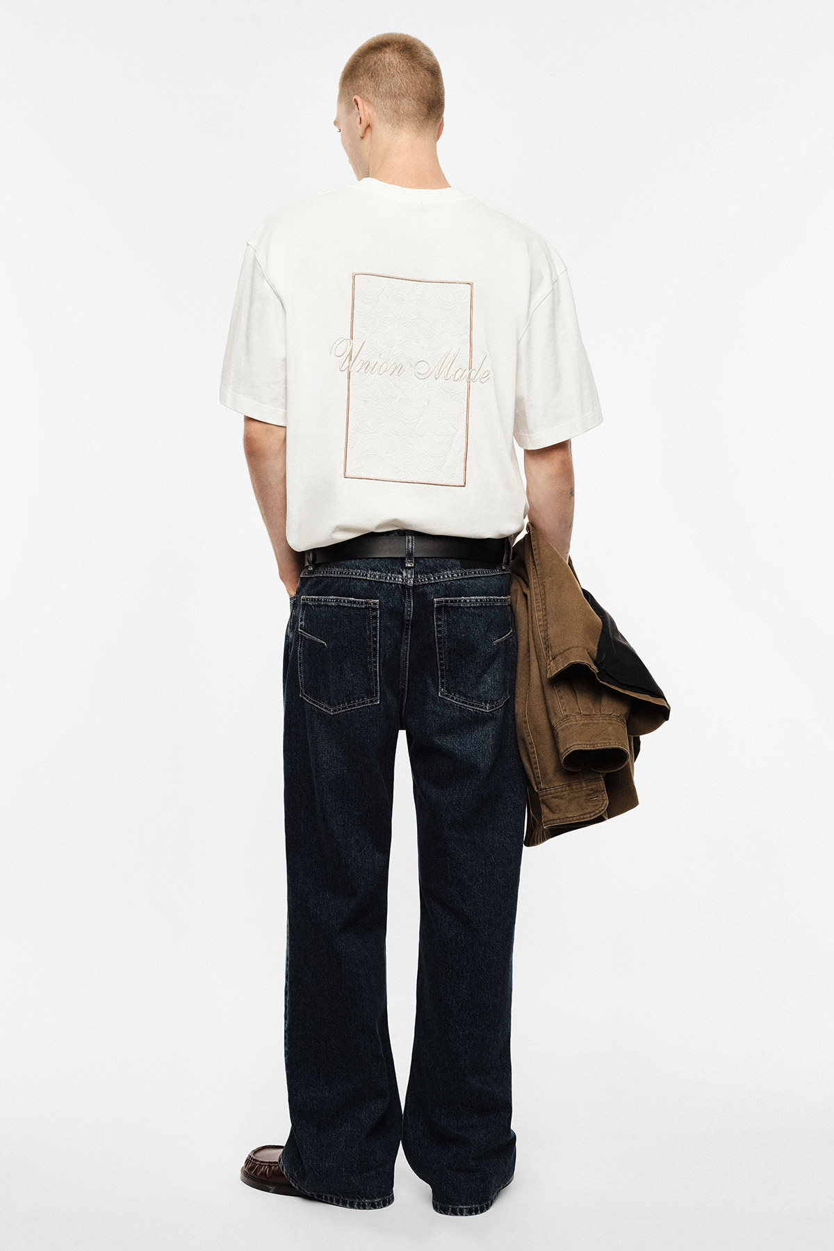 Person seen from back wearing a white graphic T‑shirt with rectangular design, dark wide‑leg jeans and holding a brown jacket