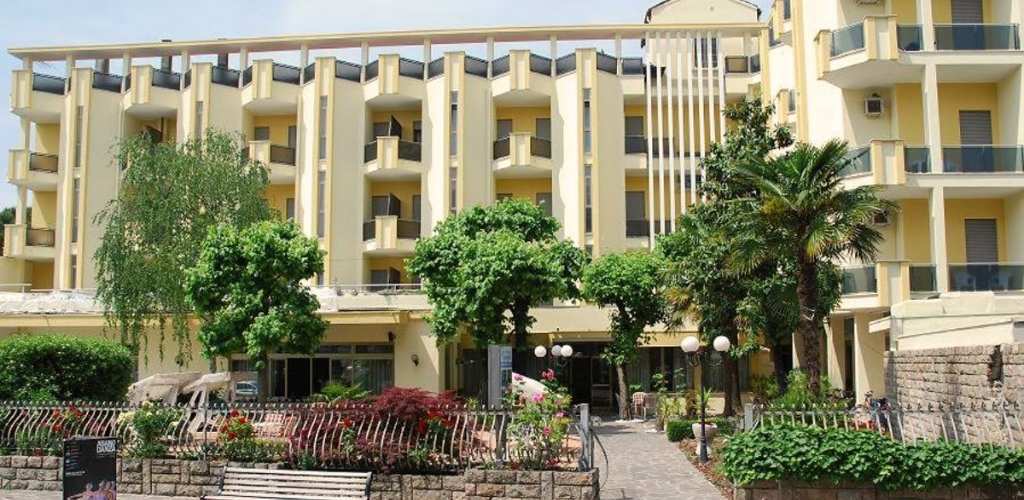 Yellow hotel building with multiple balconies, landscaped front garden, palm tree and paved entrance path