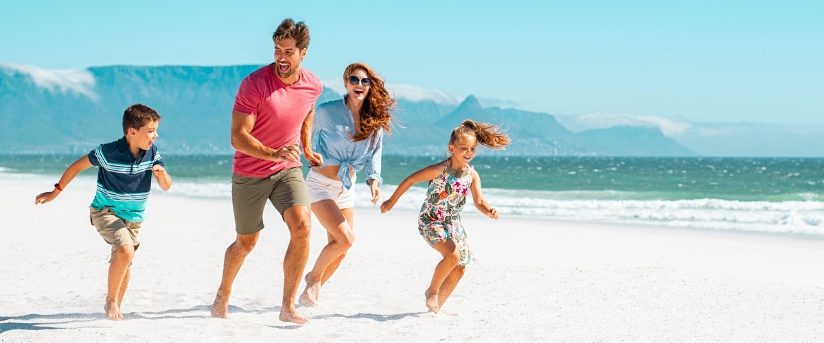 Family of four (parents and two children) running barefoot along a sunny sandy beach with turquoise sea and distant mountains