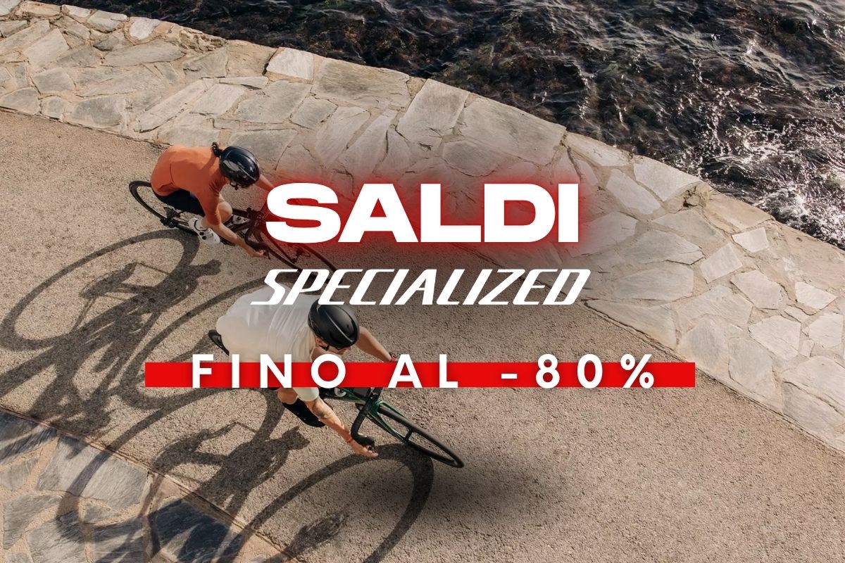 Overhead view of two cyclists on a coastal paved path beside the sea with promotional text 'SALDI Specialized fino al -80% (sale up to 80%)'