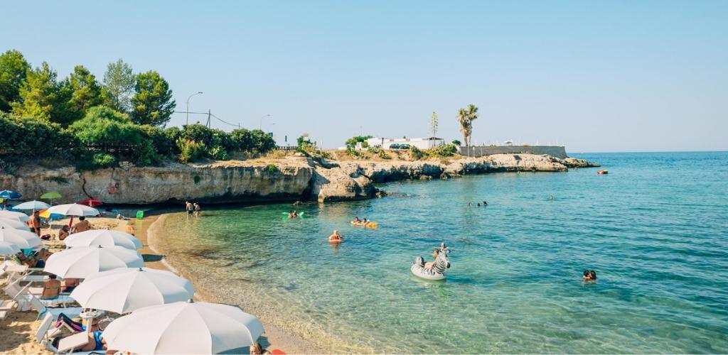 Sunny Mediterranean cove beach with white umbrellas, sunbathers and swimmers in clear turquoise water near rocky cliffs and an inflatable zebra
