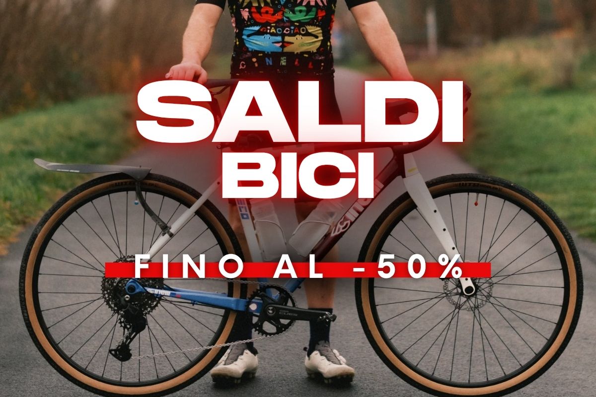 Person standing behind a gravel bike on a rural road with large 'SALDI BICI' overlay and 'FINO AL -50%' sale banner