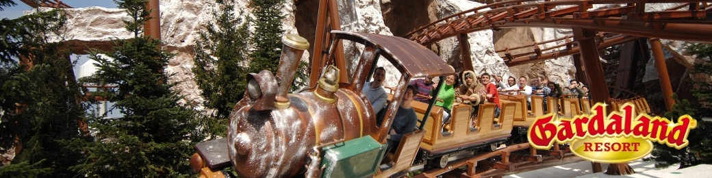 Passengers enjoying a wooden mountain-themed rollercoaster at Gardaland Resort, train emerging from a rocky tunnel beneath conifers