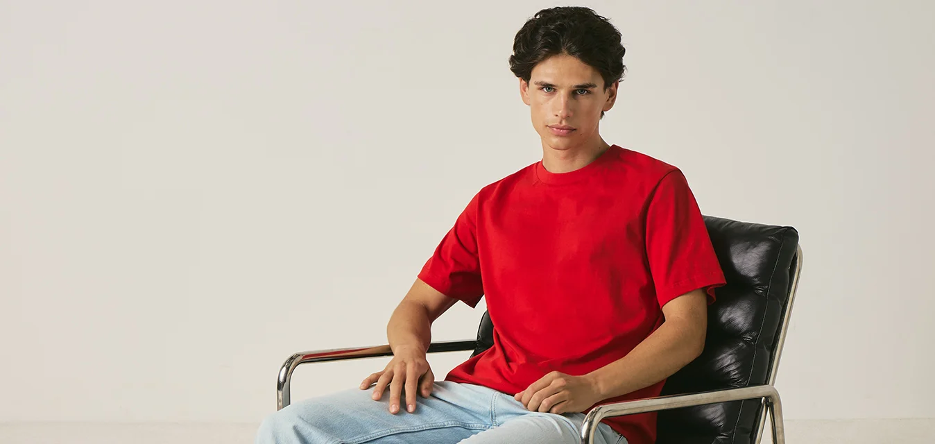 Giovane uomo con camicia rossa e jeans chiari seduto su una sedia in pelle nera con braccioli in metallo contro uno sfondo chiaro e uniforme.