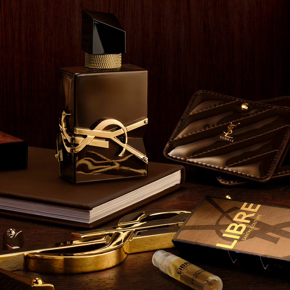 Flacone di profumo LIBRE SANTAL COUTURE su un tavolo in legno scuro accanto al suo campione, uno specchietto da borsa in pelle marrone trapuntata e accessori dorati YSL.