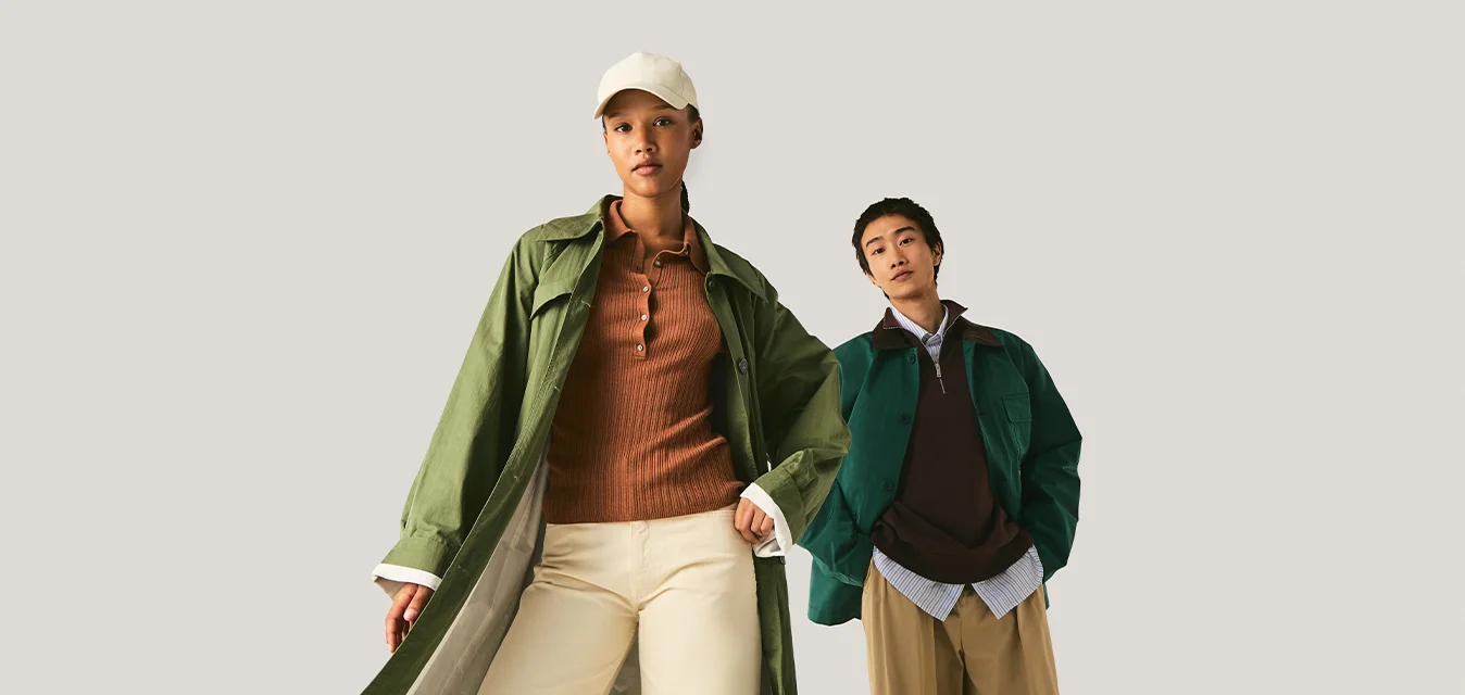 Due modelli indossano abbigliamento autunnale a strati, inclusi cappotti verdi, top marroni, pantaloni beige e color sabbia a gamba larga su uno sfondo chiaro e uniforme.