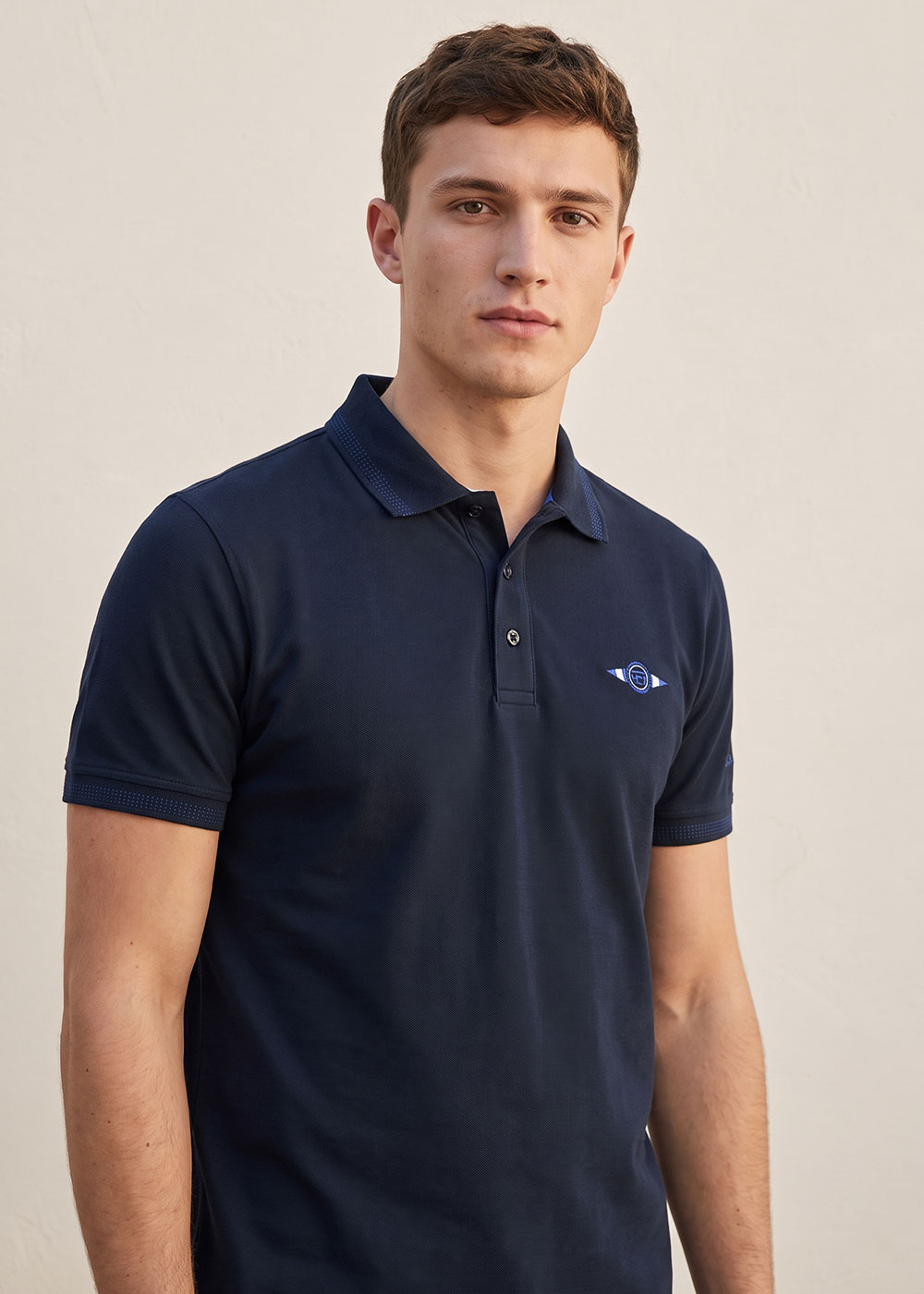 Young man modelling navy polo shirt with small embroidered logo, short sleeves, standing against a plain light wall