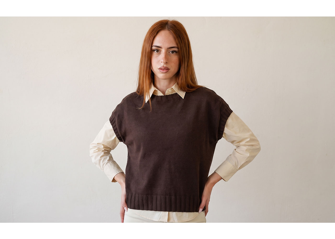 Red-haired woman in brown sleeveless jumper over cream shirt, hands on hips, neutral background portrait