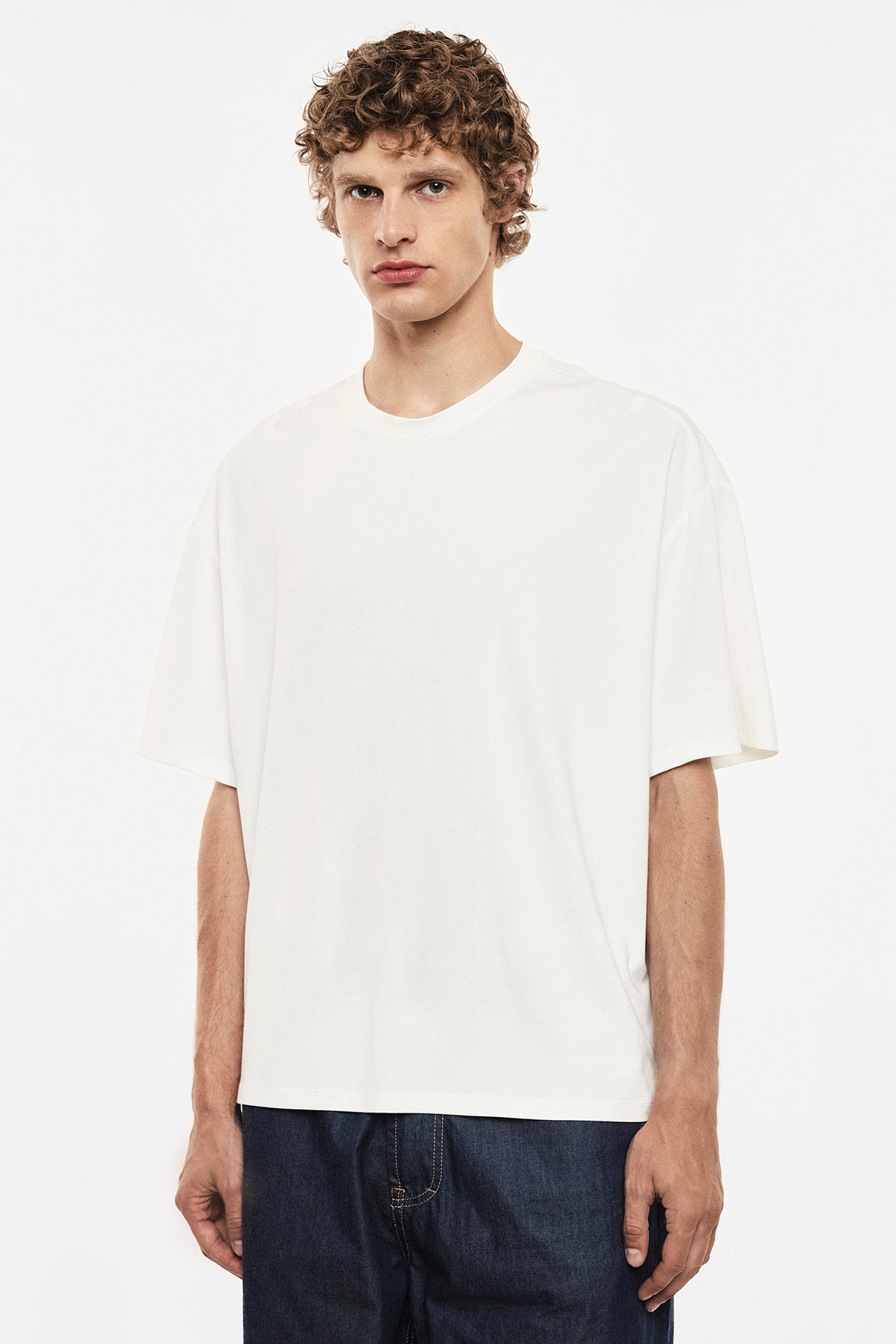 Male model wearing plain white oversized T-shirt and dark blue jeans, standing against a white studio background