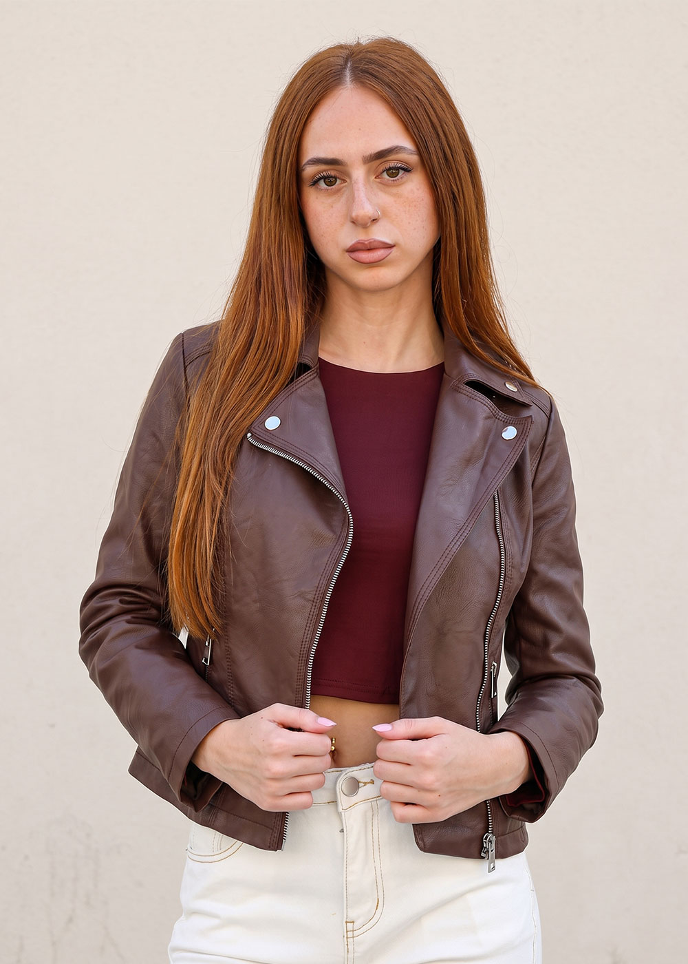 Young woman with long auburn hair wearing a brown leather jacket, maroon crop top and beige trousers against a neutral background
