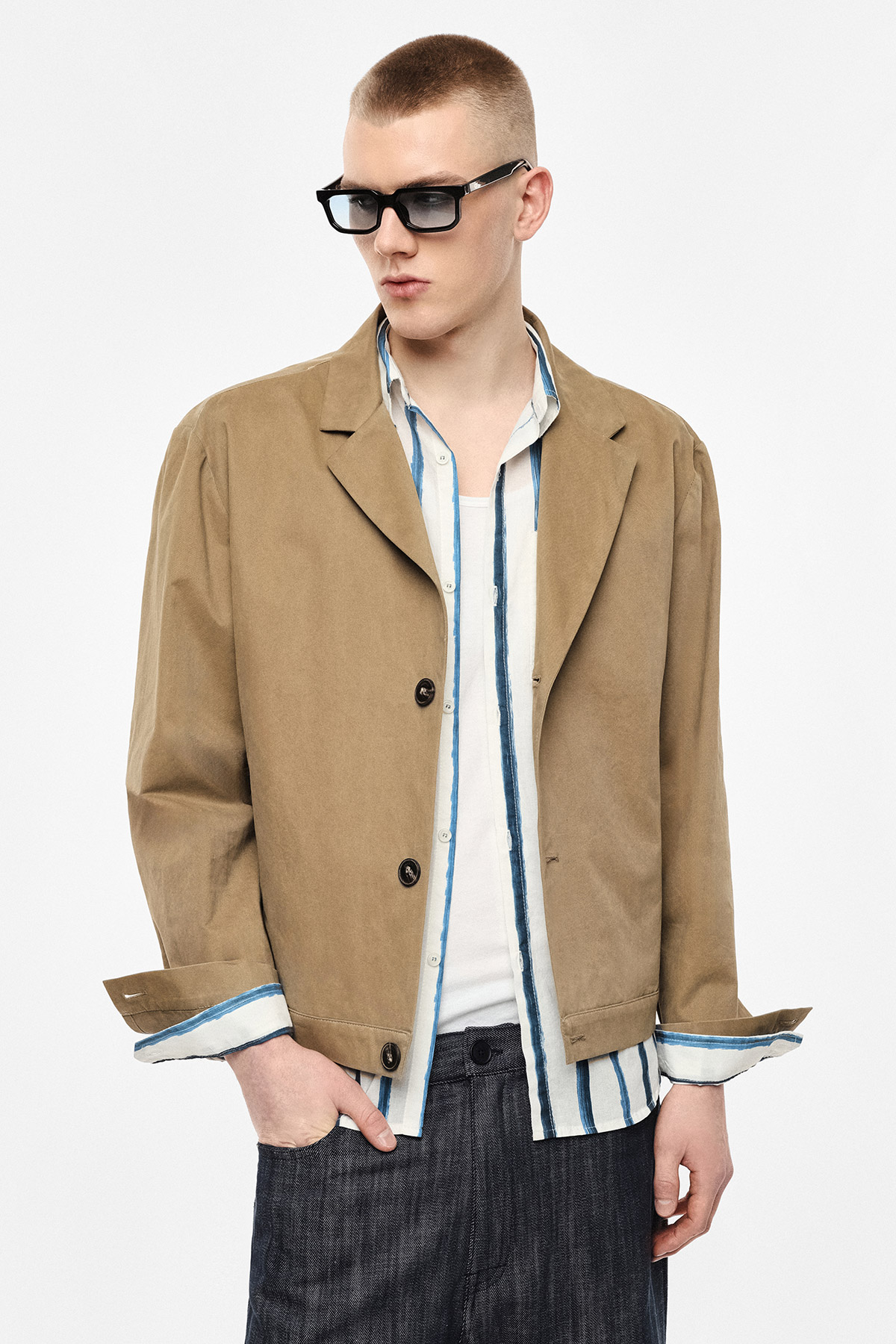 Male model wearing a beige cropped jacket over a blue-striped shirt and white vest, dark trousers and black sunglasses on white background