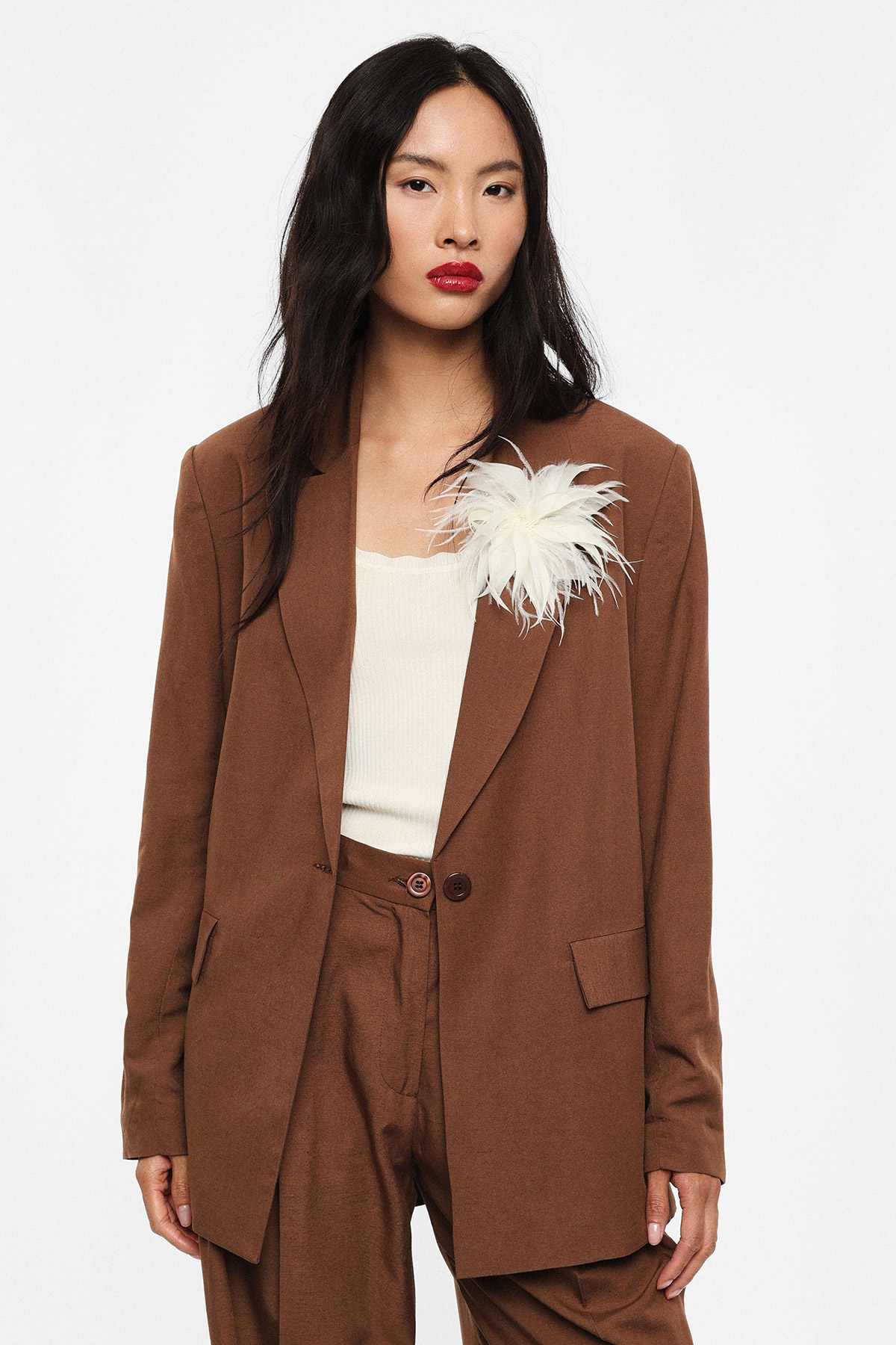 Woman wearing brown tailored blazer and trousers with cream top and white feather brooch on lapel, red lipstick