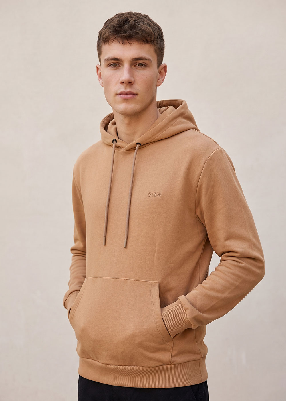 Young man wearing a camel-brown hoodie with hands in front pocket against a plain light background