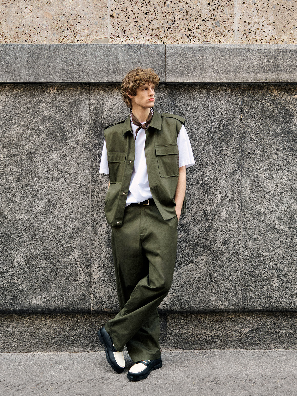Young man leaning against stone wall wearing olive-green utility vest and wide trousers, white t-shirt, neckerchief and black-and-white loafers
