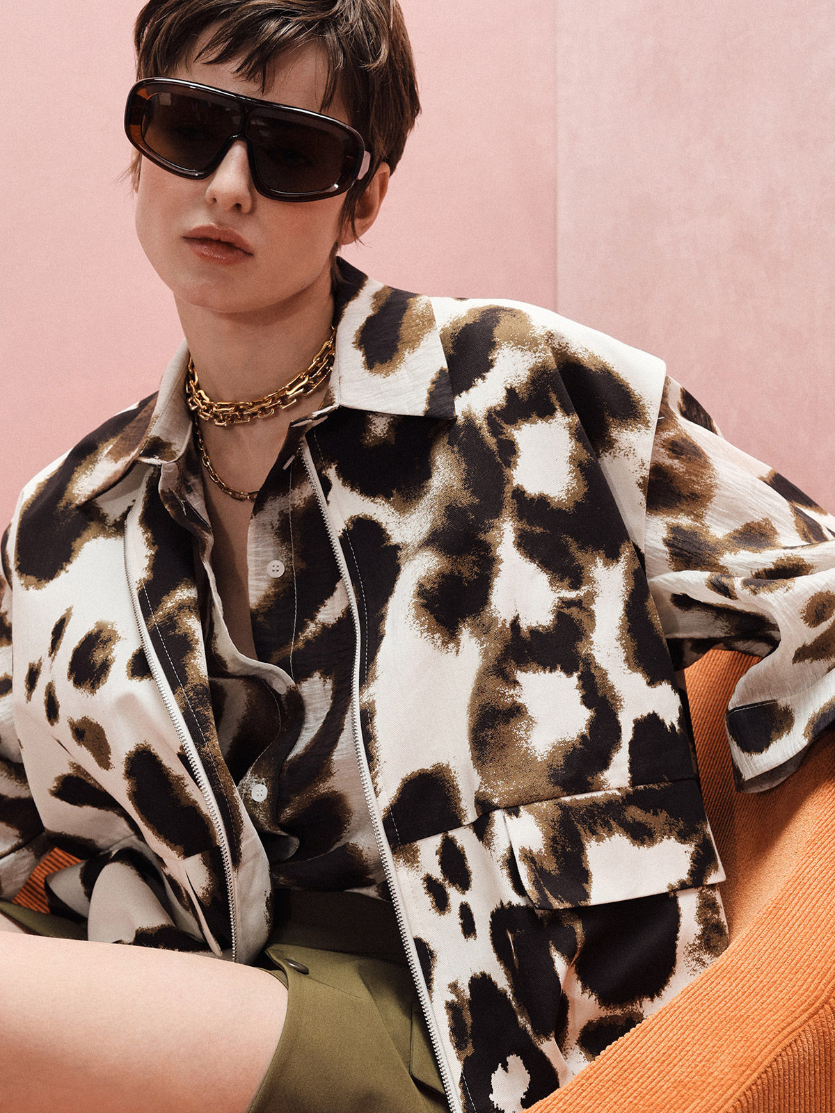 Person in oversized sunglasses, leopard-print jacket and gold chains on an orange chair against a pink background