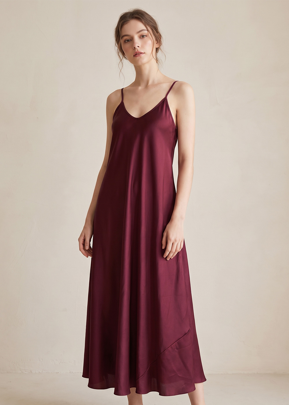 Woman wearing a burgundy satin slip dress with thin spaghetti straps and V-neck, full-length flowy gown on a neutral background