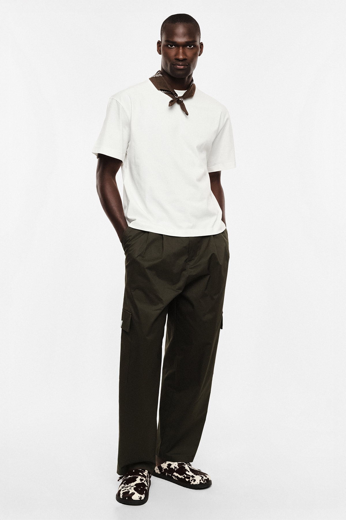 Man wearing white T-shirt, brown neck scarf, olive cargo trousers and patterned loafers, standing with hands in pockets