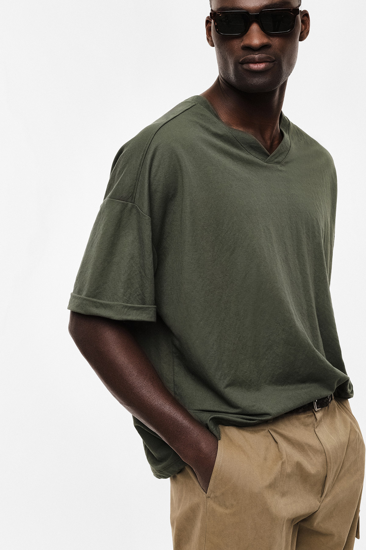 Male model wearing dark green V‑neck T-shirt, sunglasses and khaki trousers, hands in pockets on white background