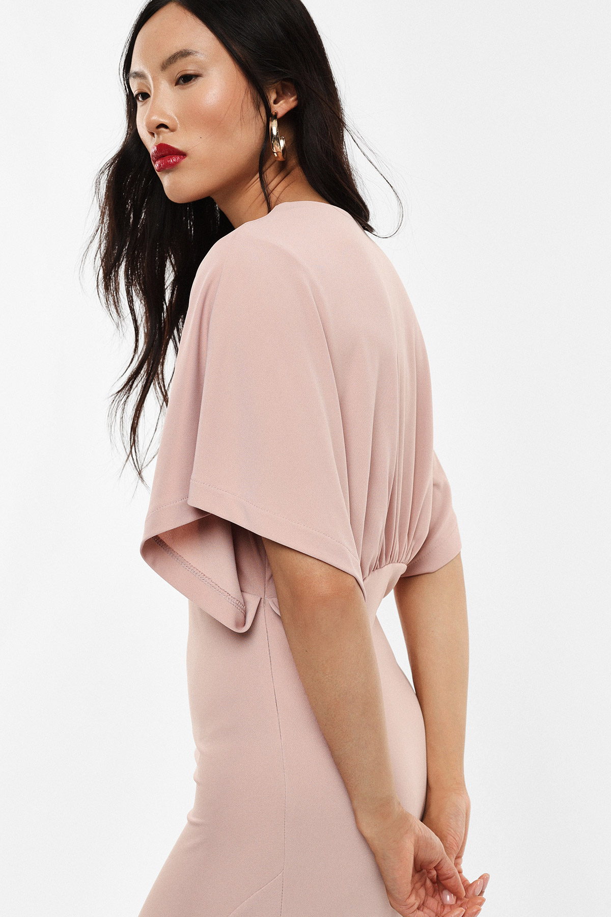 Woman in pale pink fitted dress with draped short sleeves and ruched back, side profile wearing gold hoop earrings