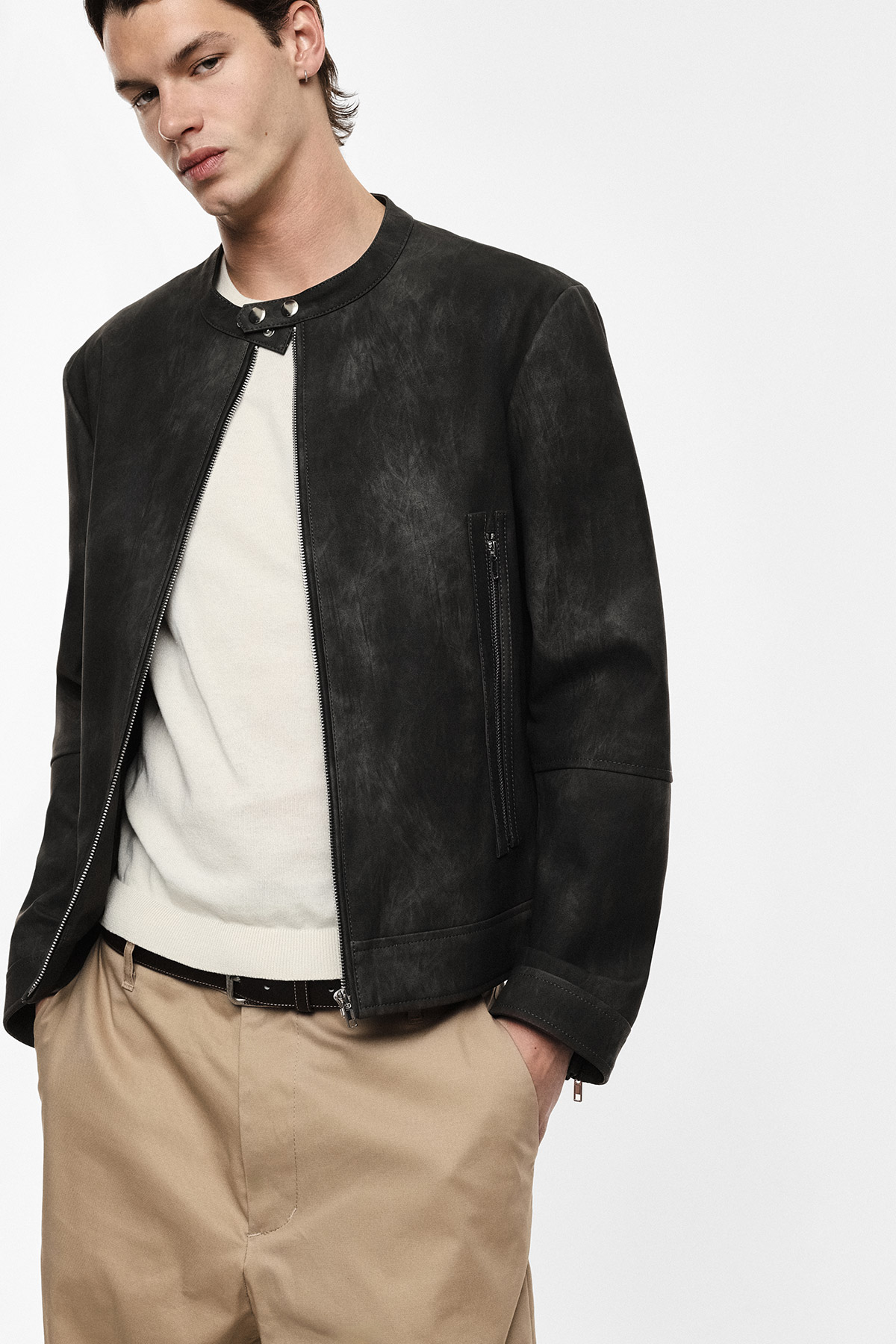 Man wearing black suede collarless biker jacket over cream sweater with beige trousers, hands in pockets against white studio background