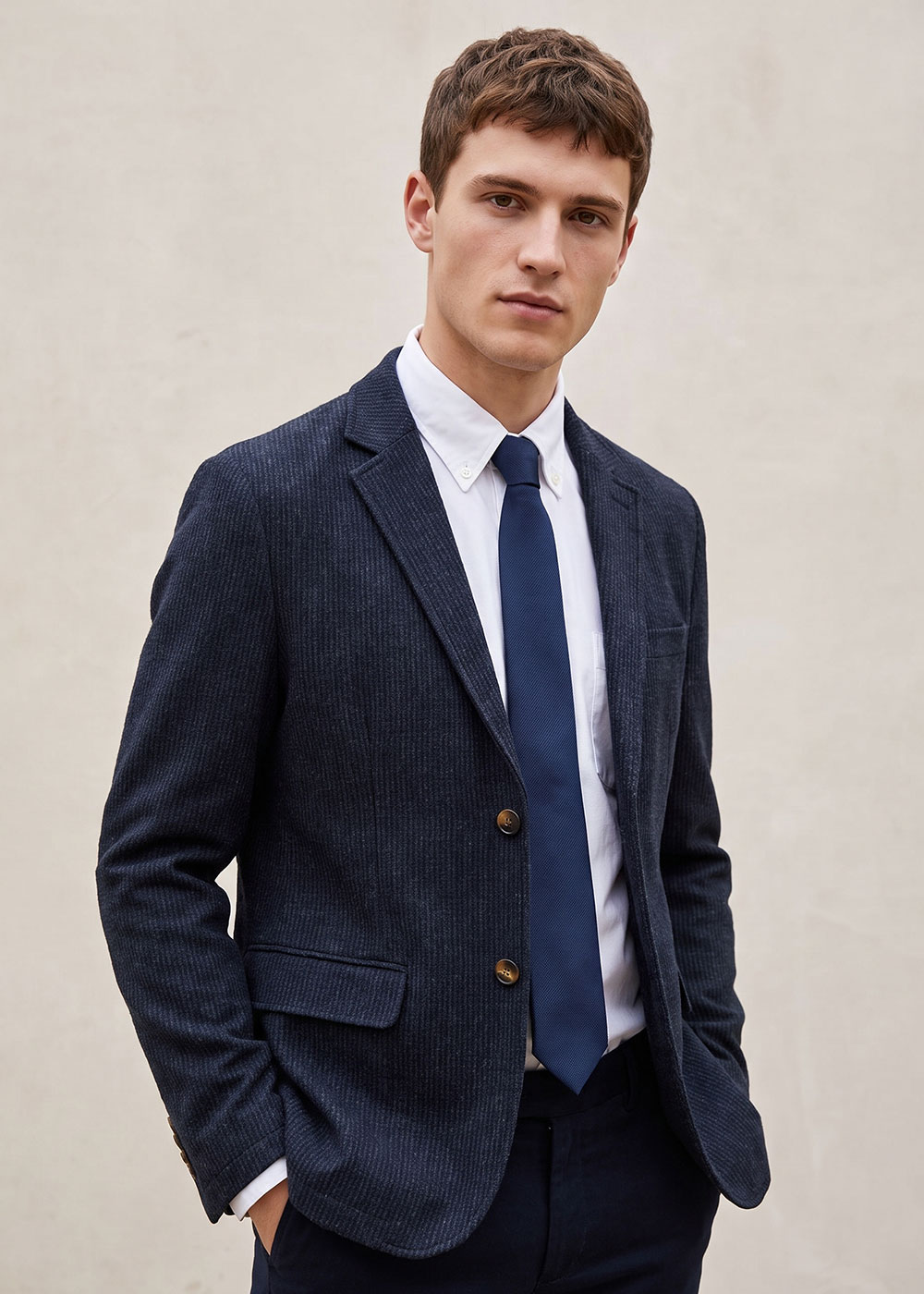 Young man wearing navy pinstripe blazer, white button-down shirt and blue tie, hands in pockets against neutral backdrop