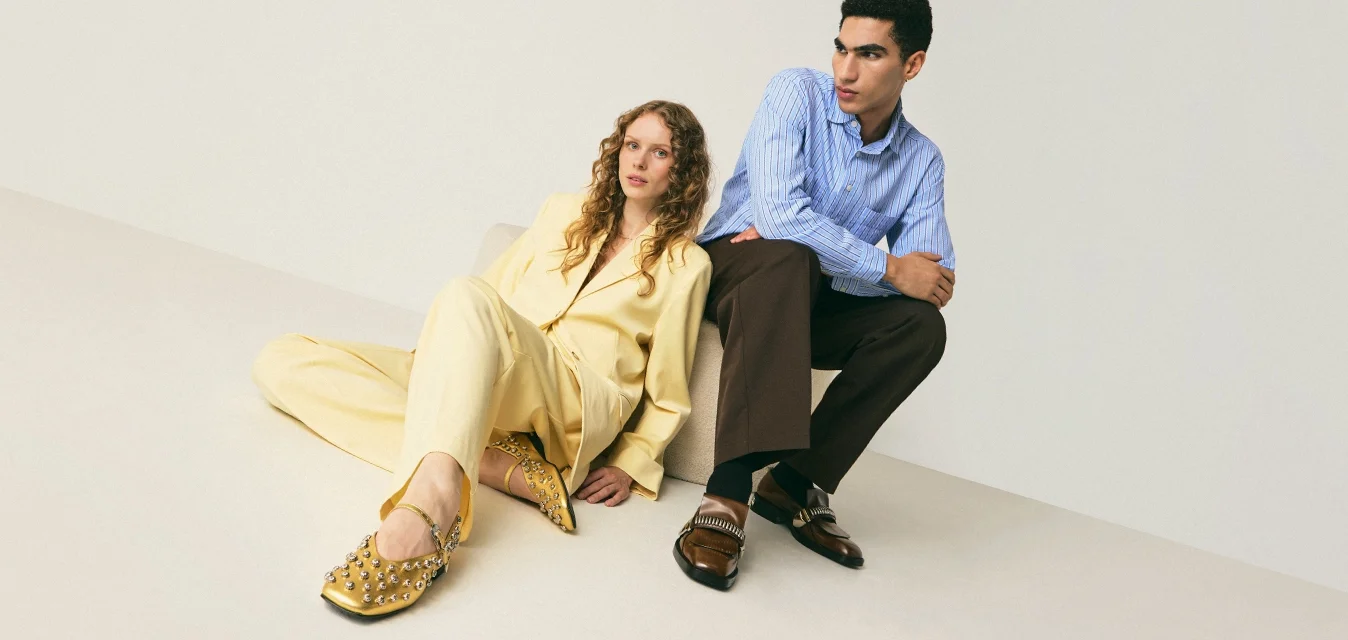Una donna in un completo giallo e scarpe con borchie è seduta per terra, un uomo in camicia blu e pantaloni scuri è seduto su un blocco accanto a lei, sullo sfondo semplice.