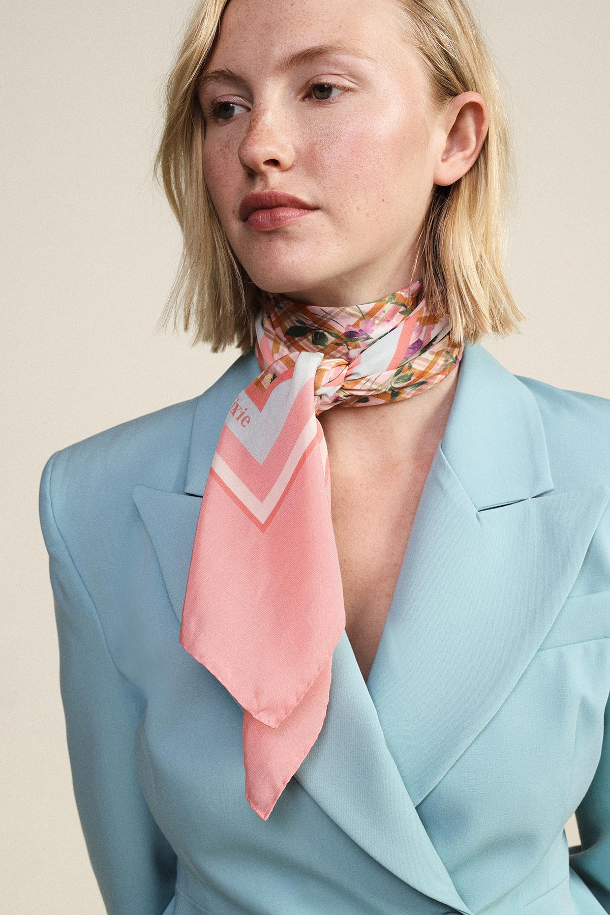 Woman in light blue blazer wearing a pink patterned silk neck scarf, short blonde hair, neutral background