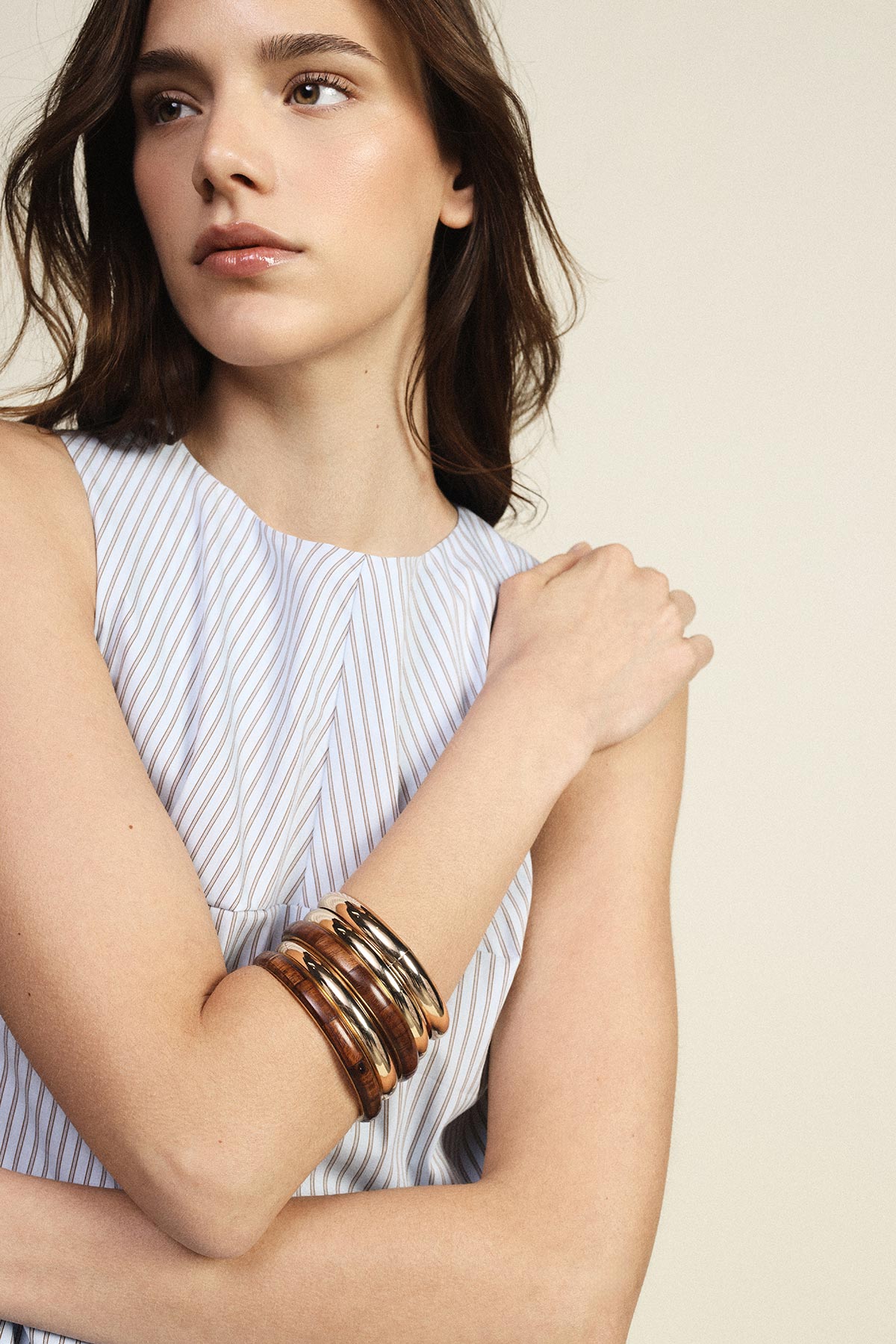 Close-up of young woman in sleeveless striped dress wearing stacked metallic bangles on her forearm, looking sideways against a neutral background
