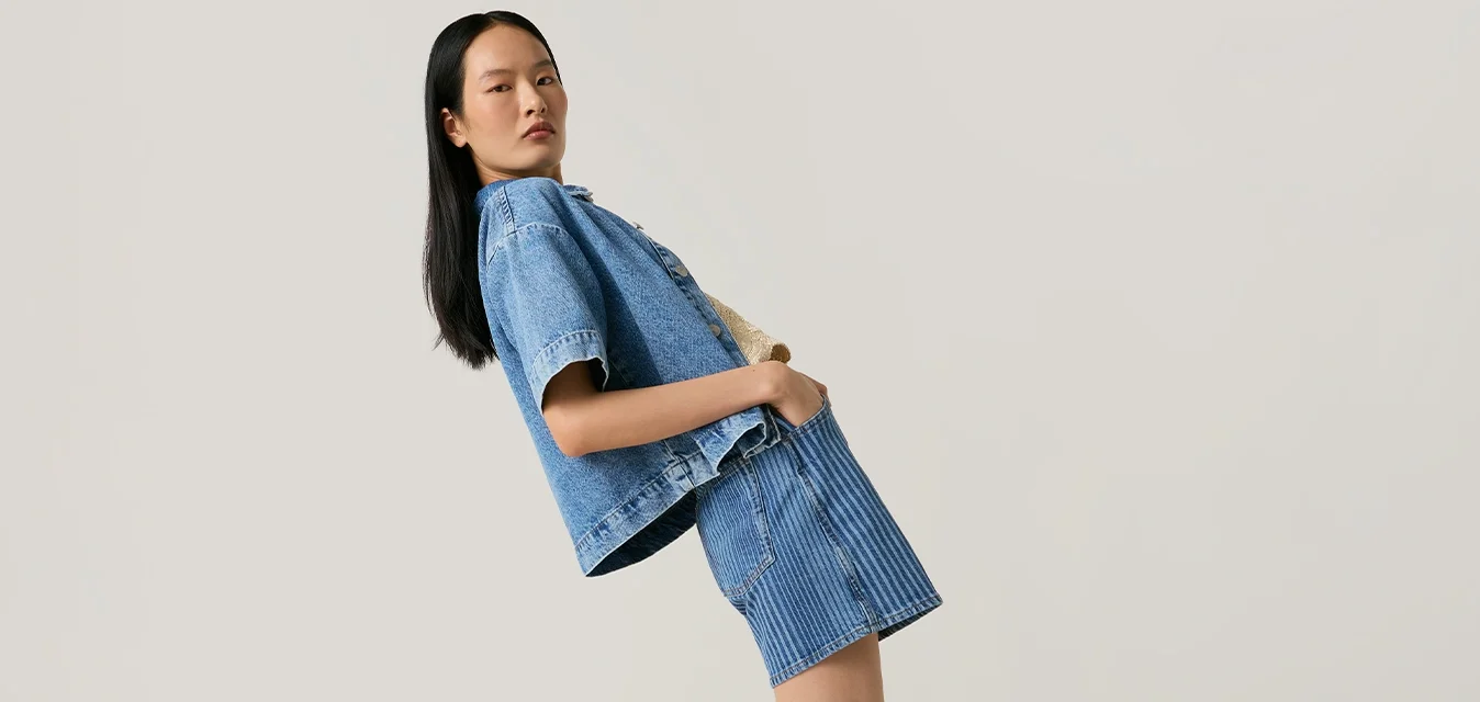 Giovane donna che si piega all'indietro indossando una giacca di jeans e una gonna con stivali da cowboy marroni su uno sfondo chiaro e uniforme.