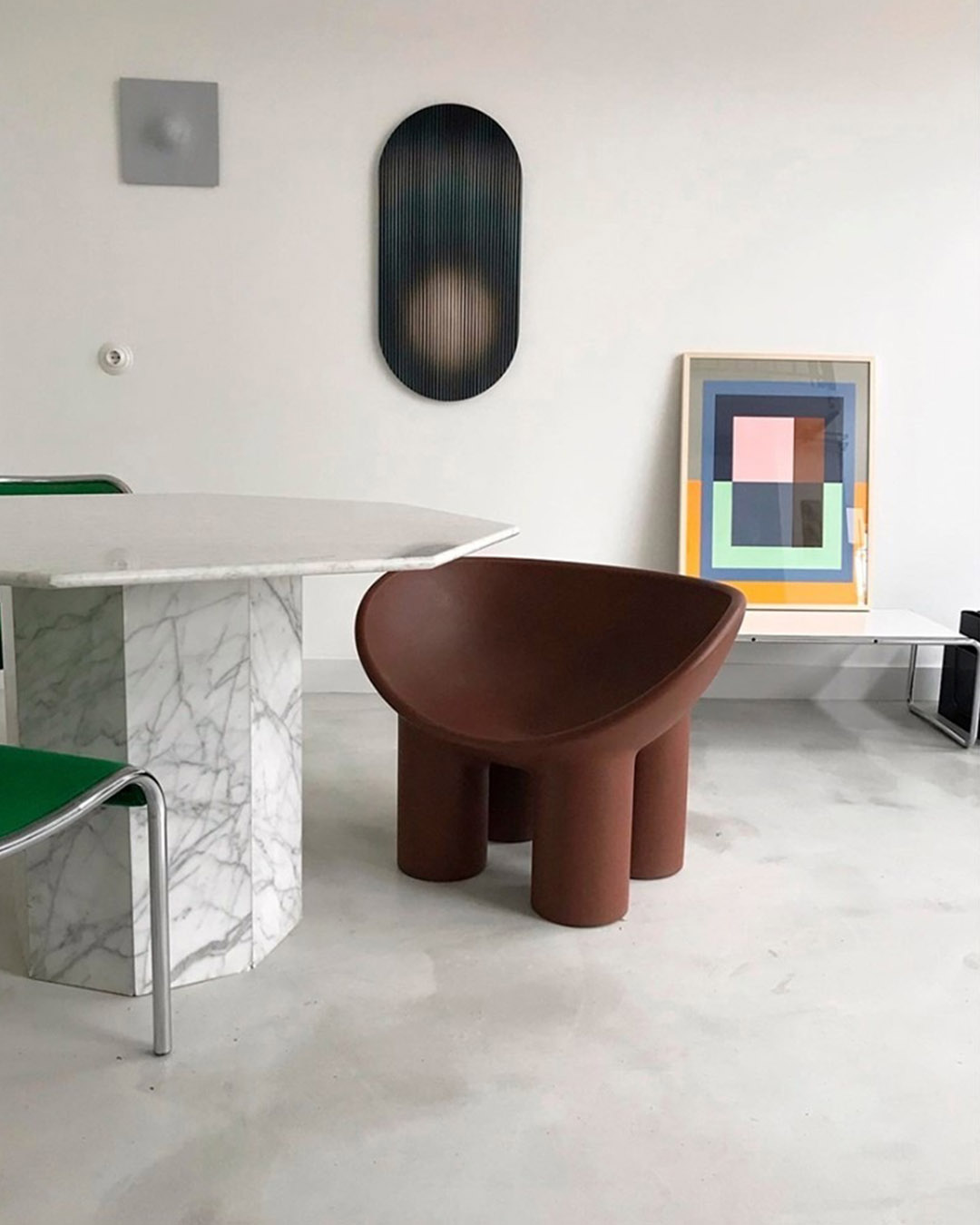 Minimalist interior with brown sculptural chair on cylindrical legs beside marble dining table and colourful abstract artwork