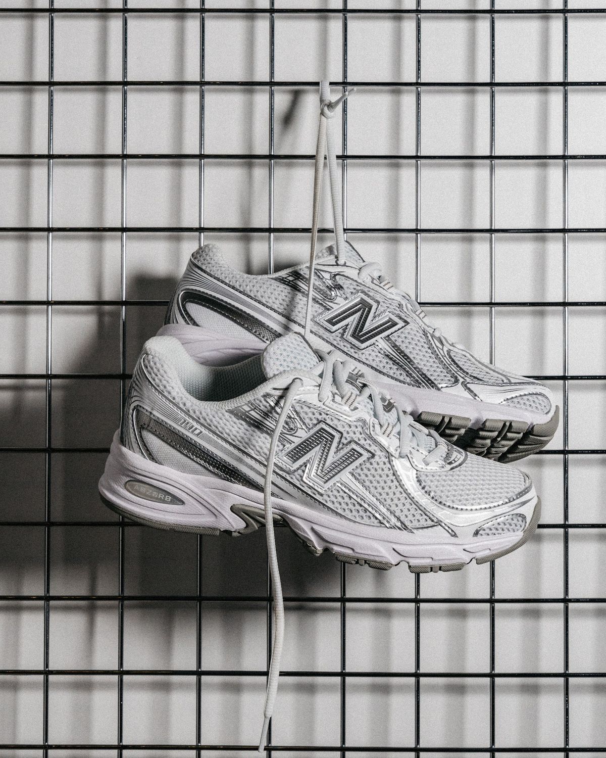 Pair of white and silver New Balance running trainers hanging by their laces on a metal grid display
