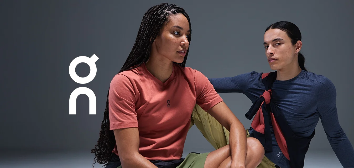 Due persone siedono su uno sfondo chiaro e uniforme. Una indossa una maglietta corallo con pantaloni corti verdi, l'altra una maglia a maniche lunghe navy con pantaloni corti simili.