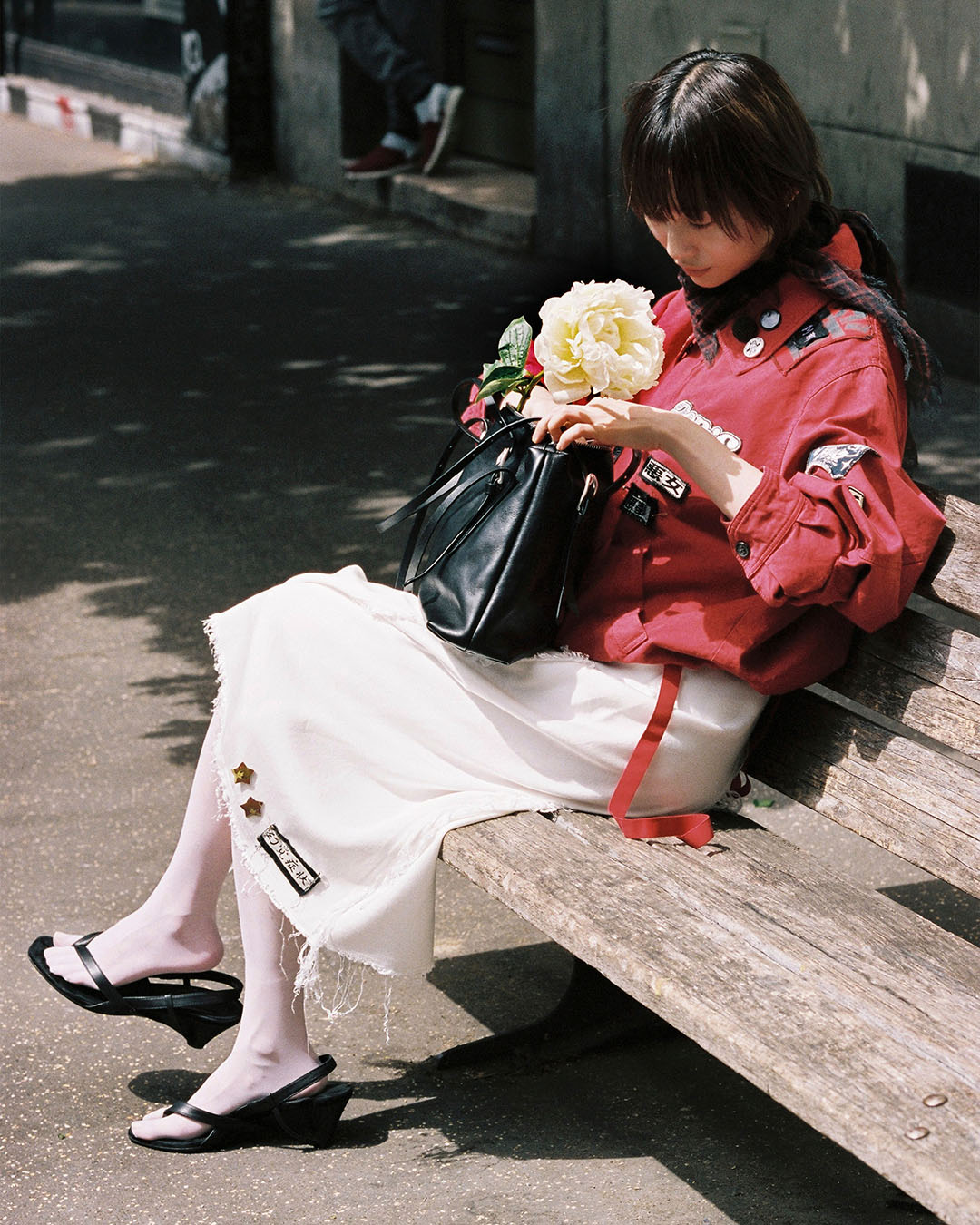 Young woman in red jacket and white skirt sitting on park bench holding a peony and black handbag
