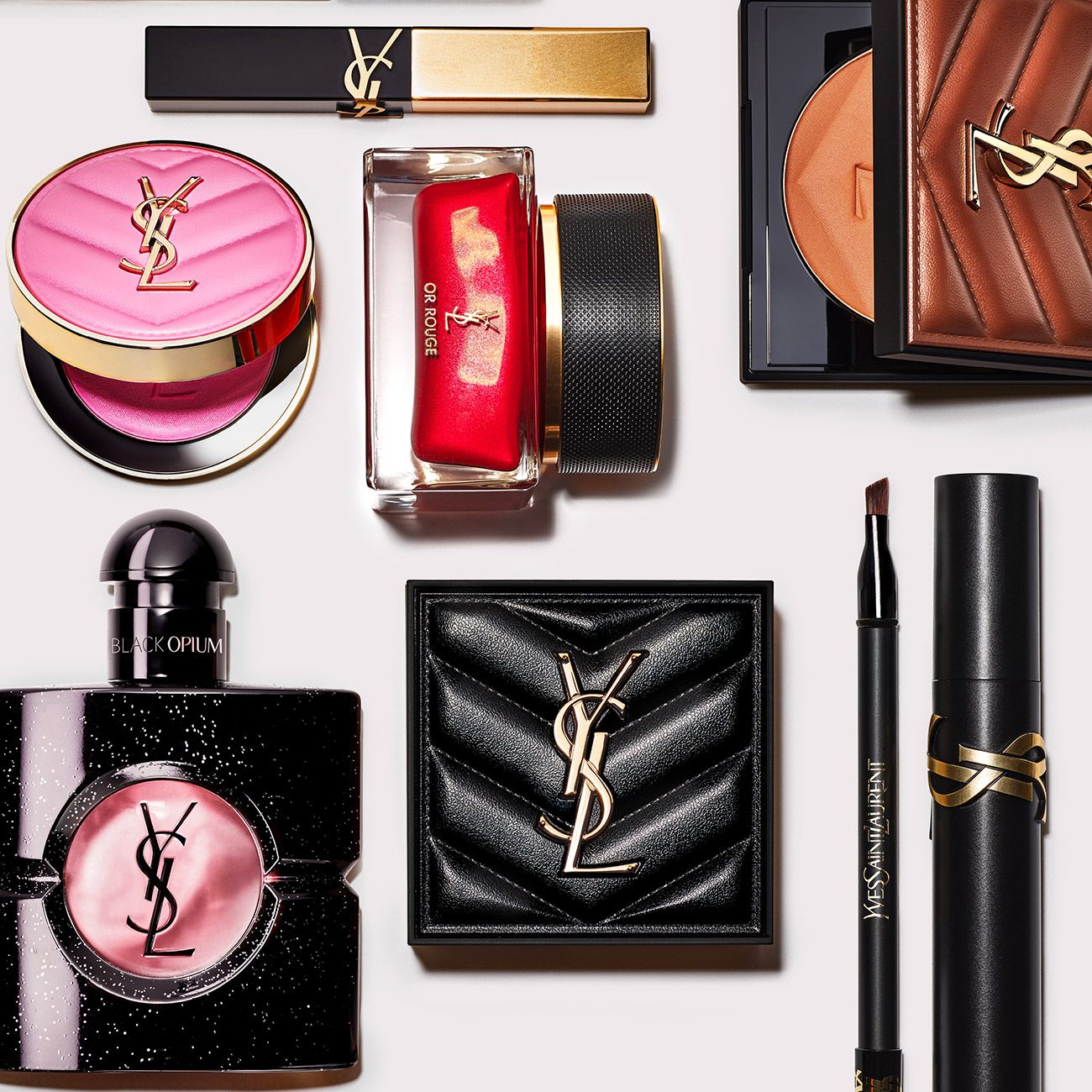 Una selezione di prodotti da trucco YSL e un flacone di profumo Black Opium disposti dall'alto su sfondo chiaro.