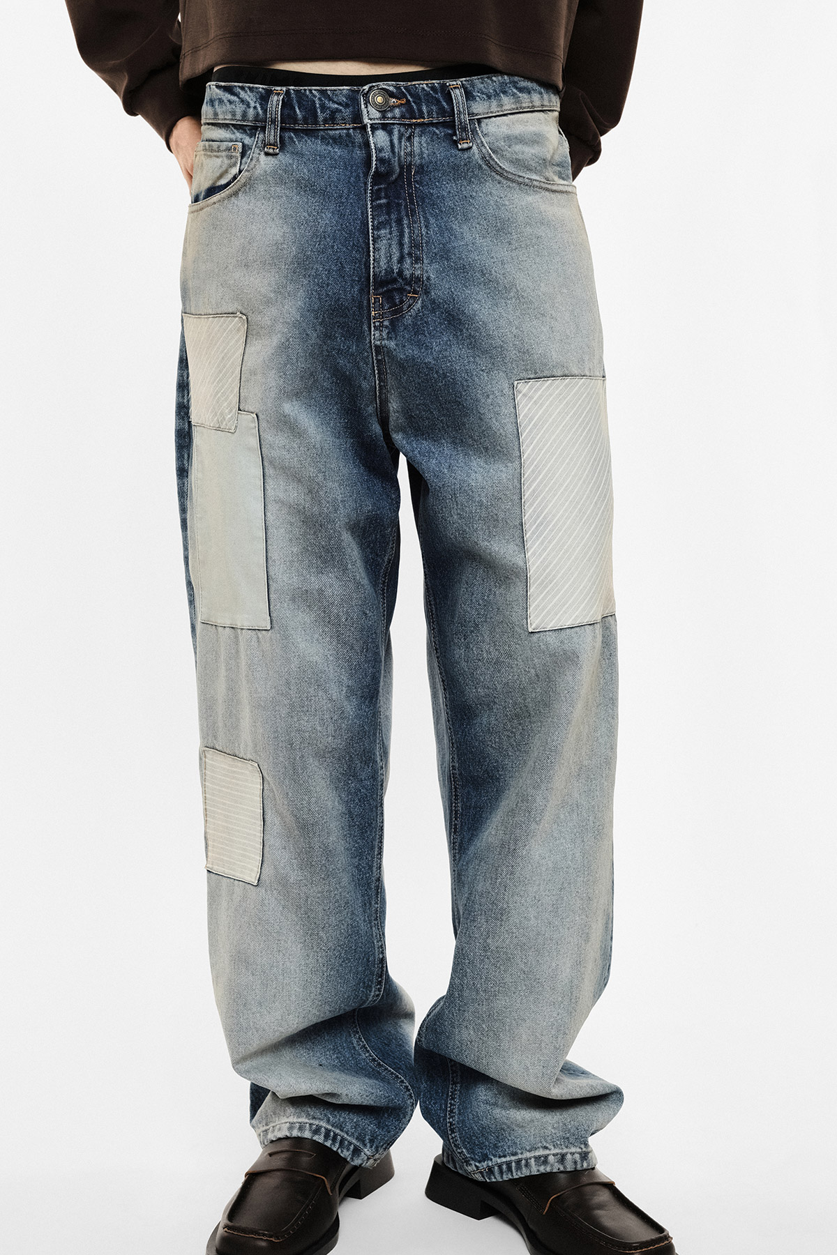 Men's faded blue relaxed baggy jeans with light rectangular patchwork on front and sides, paired with black leather loafers