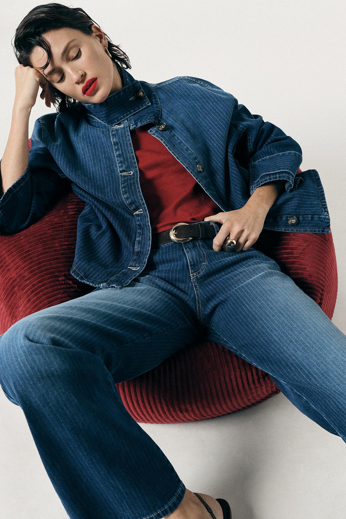 Woman reclining in a red chair wearing denim jacket and jeans with red top and black belt