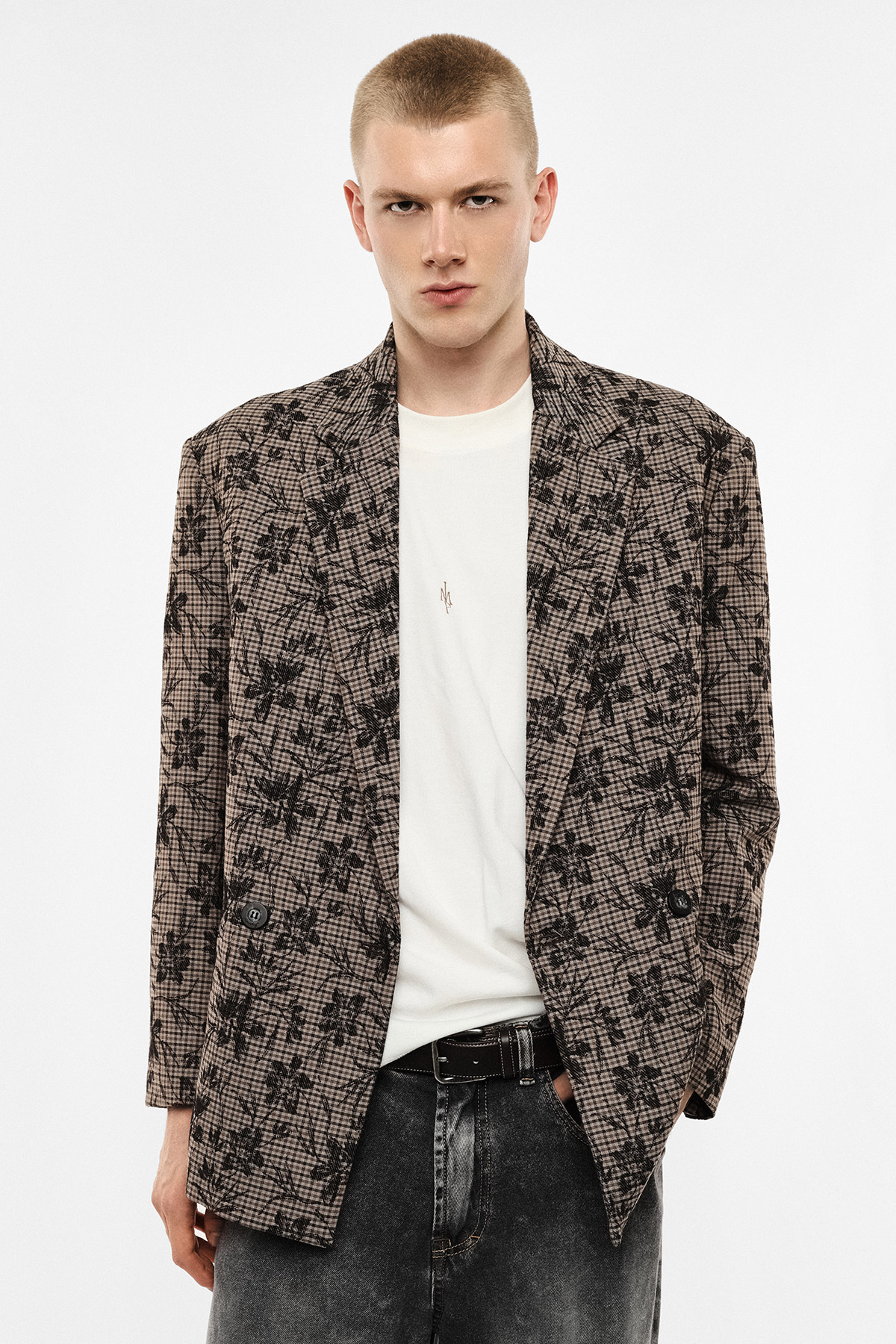 Young man wearing brown checked floral blazer over white T-shirt and black jeans, standing against a plain light background