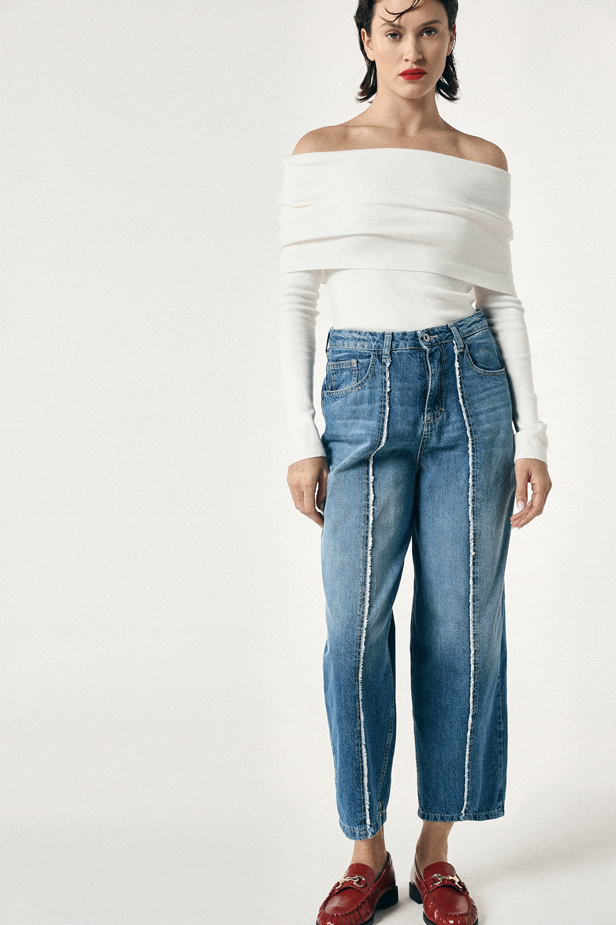 Woman wearing high‑waist cropped wide‑leg blue jeans with frayed vertical seams, paired with white off‑shoulder top and red loafers