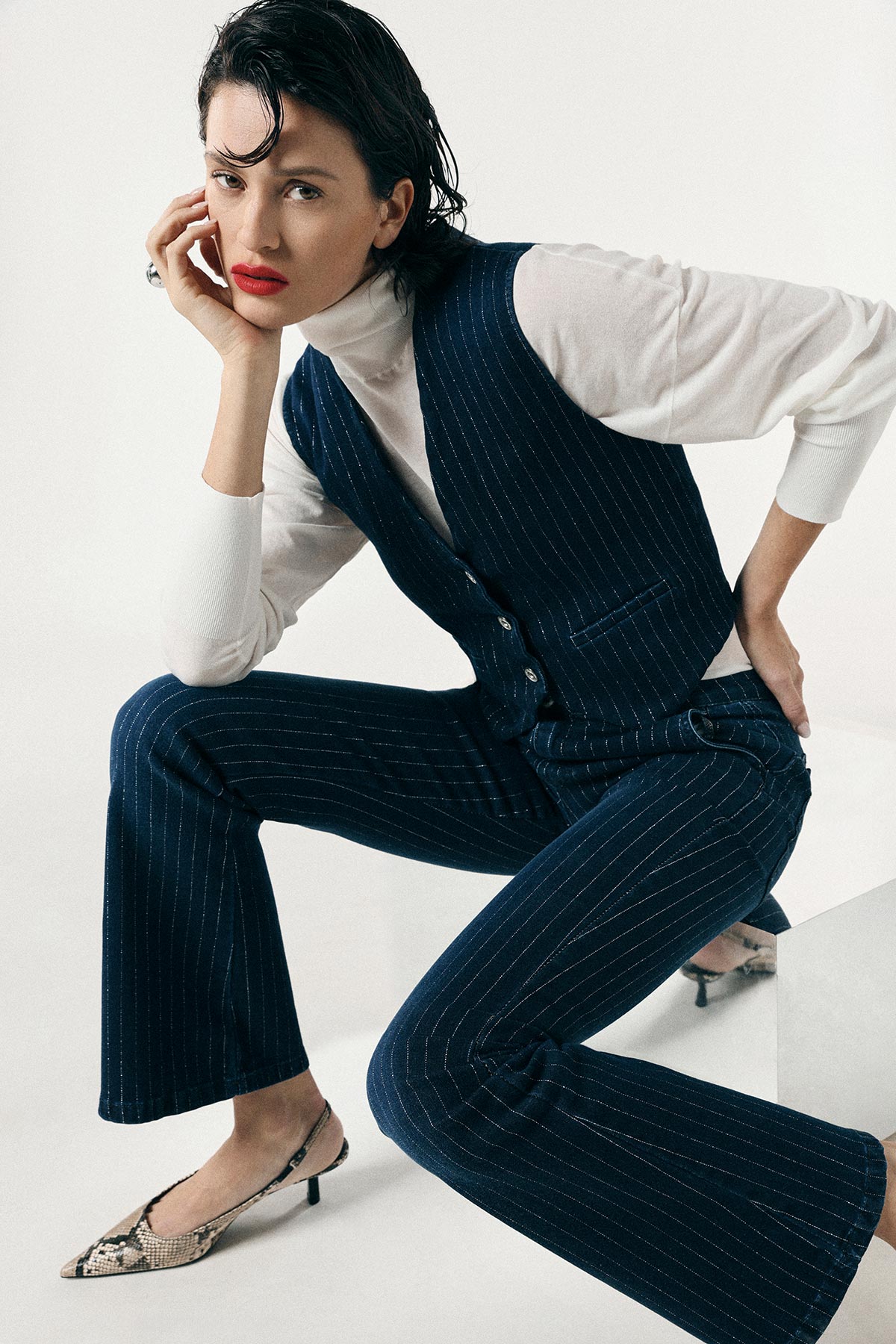 Woman in navy pinstripe waistcoat and trousers with white roll‑neck, red lipstick, seated pose, snakeskin heels