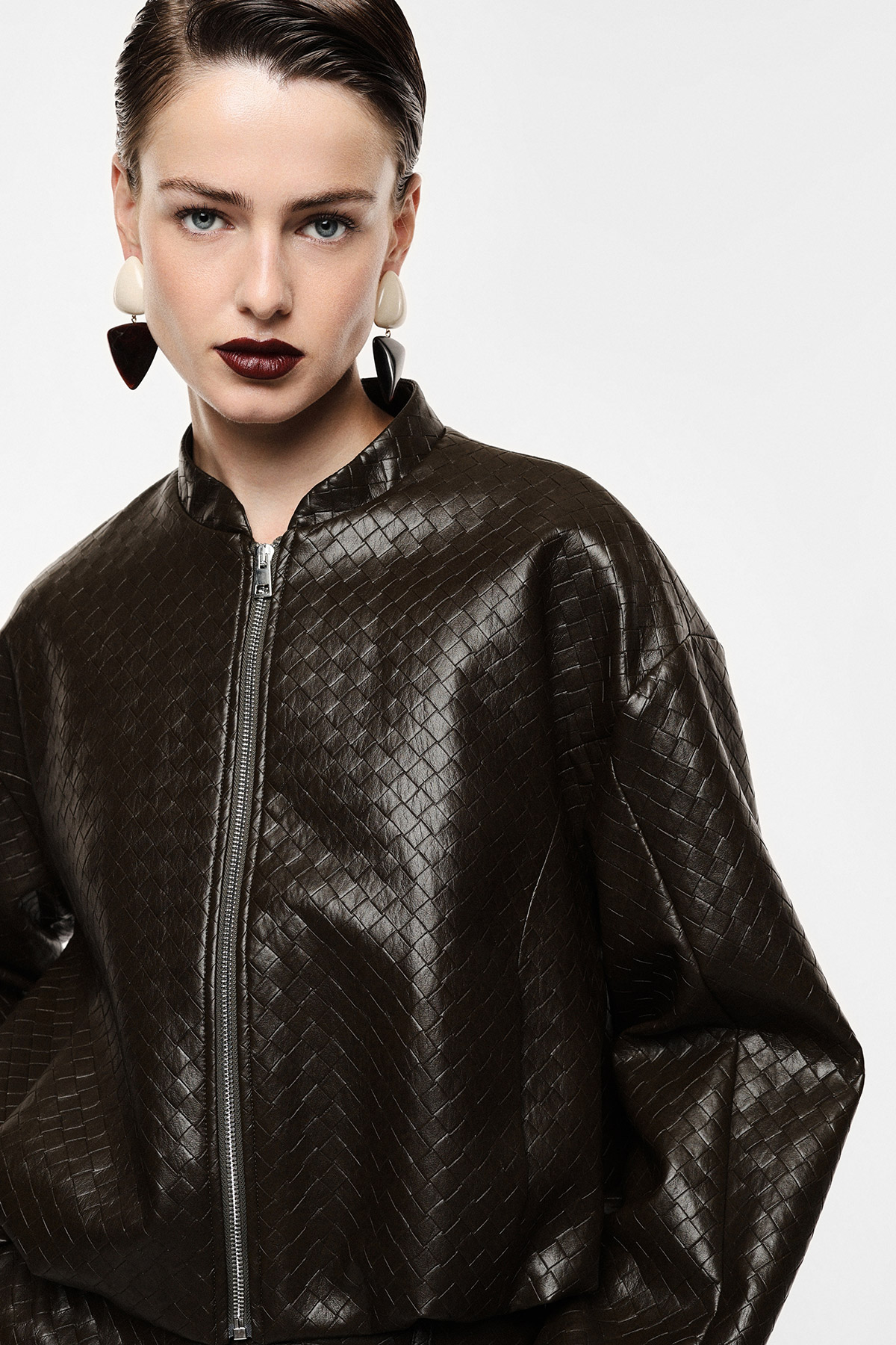 Female model in a dark brown textured leather zip jacket with bold geometric earrings and glossy burgundy lipstick, studio portrait