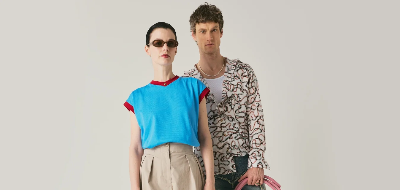 Due modelli stanno in piedi con tubi rosa arrotolati; la donna indossa un top blu, culottes beige, occhiali da sole e tiene una borsa gialla con borchie; l'uomo indossa una camicia a fantasia e pantaloncini di jeans.