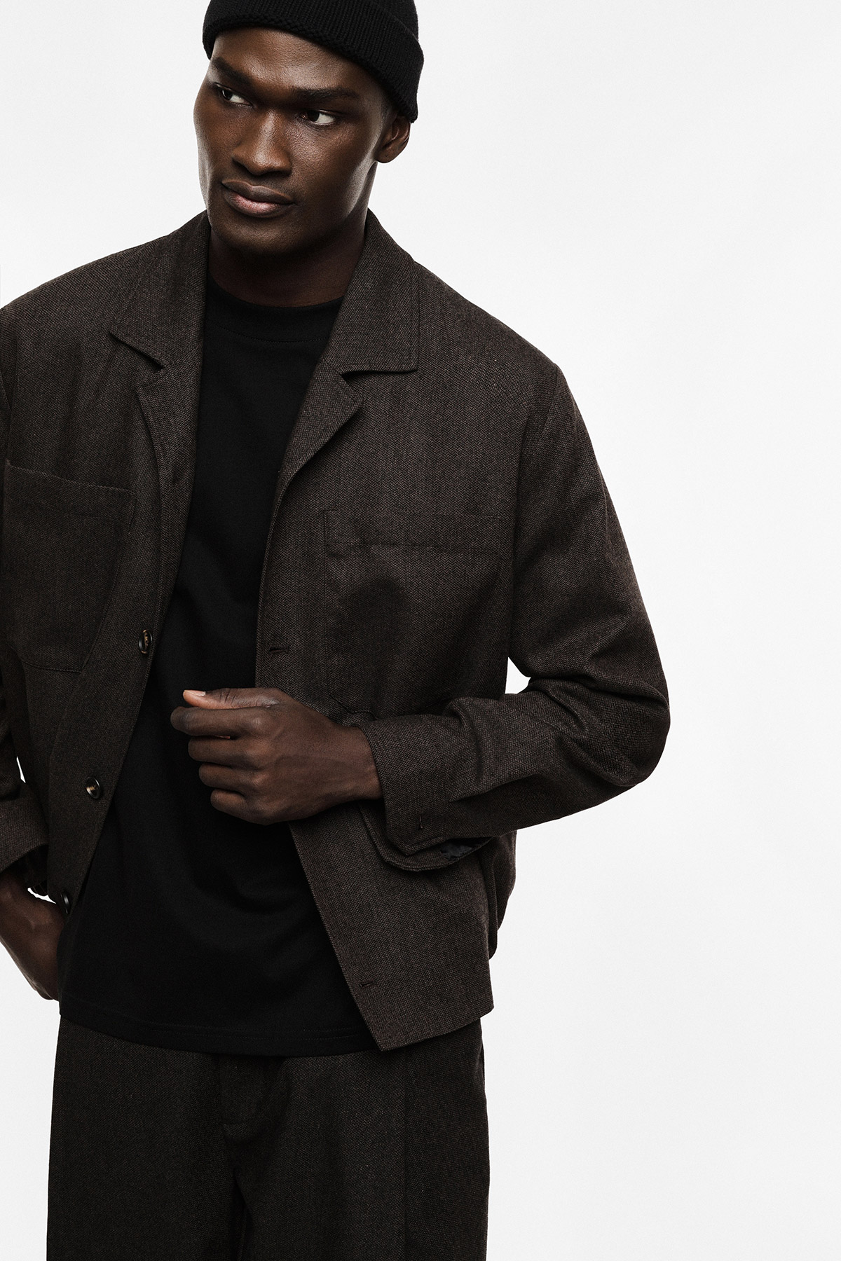 Man in brown textured jacket, black T-shirt and black beanie in a cropped menswear fashion portrait on white background