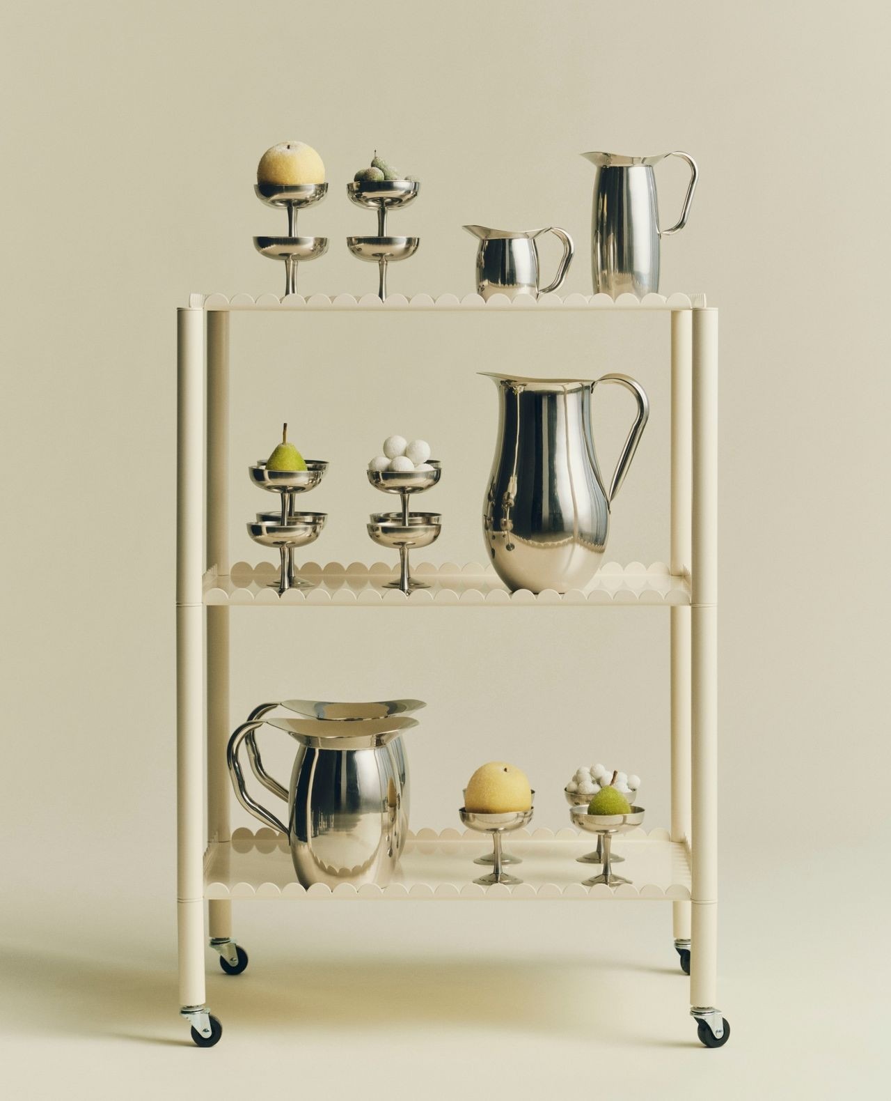 Cream metal serving trolley with three shelves displaying polished silver pitchers and tiered bowls of fruit and small white spheres