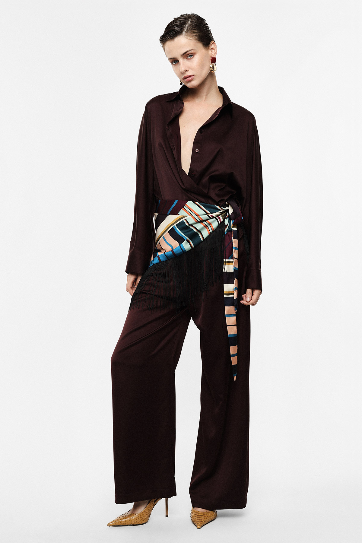Model in dark brown wrap-style shirt and wide-leg trousers, colourful striped fringed sash, gold heels and statement earring