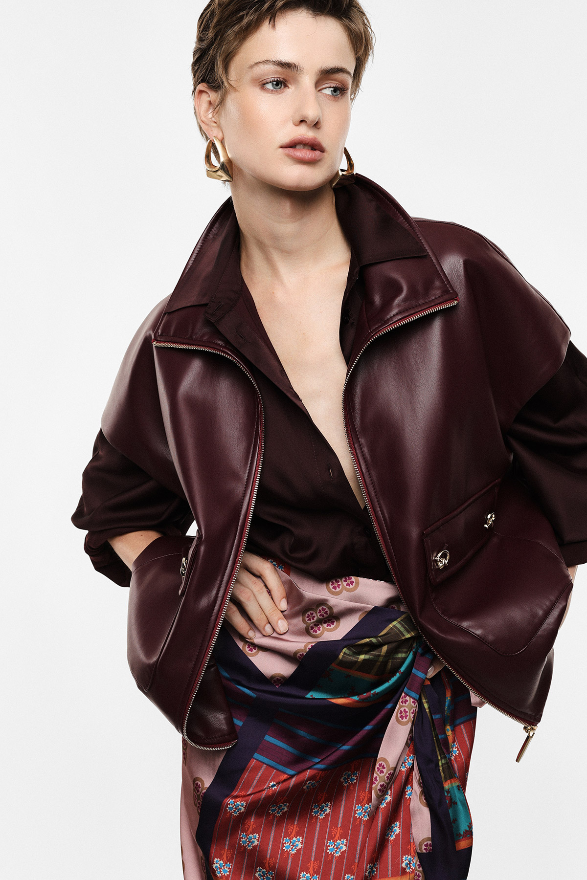 Woman modelling burgundy leather jacket, dark blouse, patterned silk wrap skirt and gold hoop earrings against white background