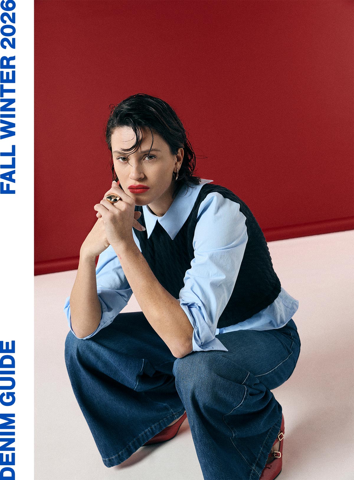 Model seated against red backdrop for Fall Winter 2026 denim guide, wearing light-blue shirt, black vest, wide-leg jeans and red heels.